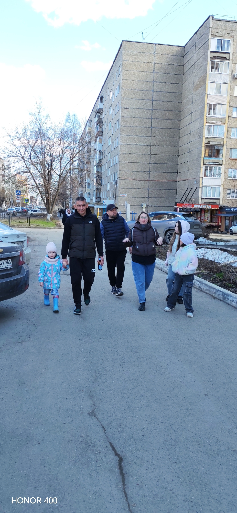 A group of six people is walking down a sidewalk in Izhevsk, Russia. A man in a black jacket and black Adidas pants is holding the hand of a small child in a blue snowsuit. Behind them are a man in a blue vest, a woman in a brown jacket and jeans, and two girls in jackets and jeans. The group is walking in front of a grey apartment building with shops on the ground floor. A dark grey car is parked to the left of the group. The text "HONOR 400" is at the bottom of the image.