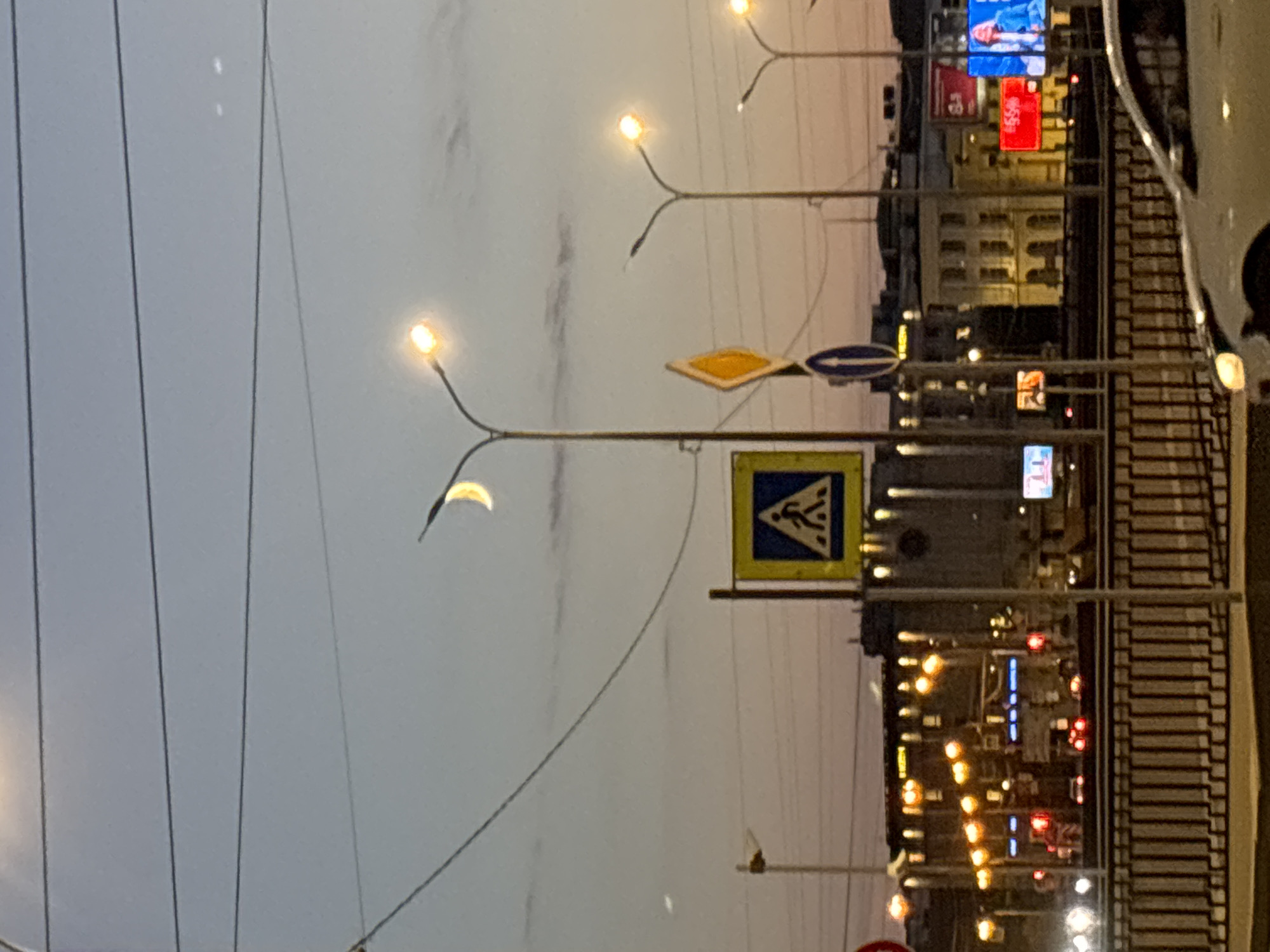 A crescent moon is visible in the twilight sky above a city street scene in Saint Petersburg, Russia. Streetlights with glowing orange bulbs illuminate the road, casting a warm light on the urban landscape. A pedestrian crossing sign, a diamond-shaped warning sign, and a blue circular sign are mounted on poles, indicating traffic regulations. In the background, buildings with lit windows form a cityscape, while billboards display advertisements. Cars are visible on the road, and a railing runs along the edge of the elevated roadway. The overall impression is of a bustling city transitioning from day to night.