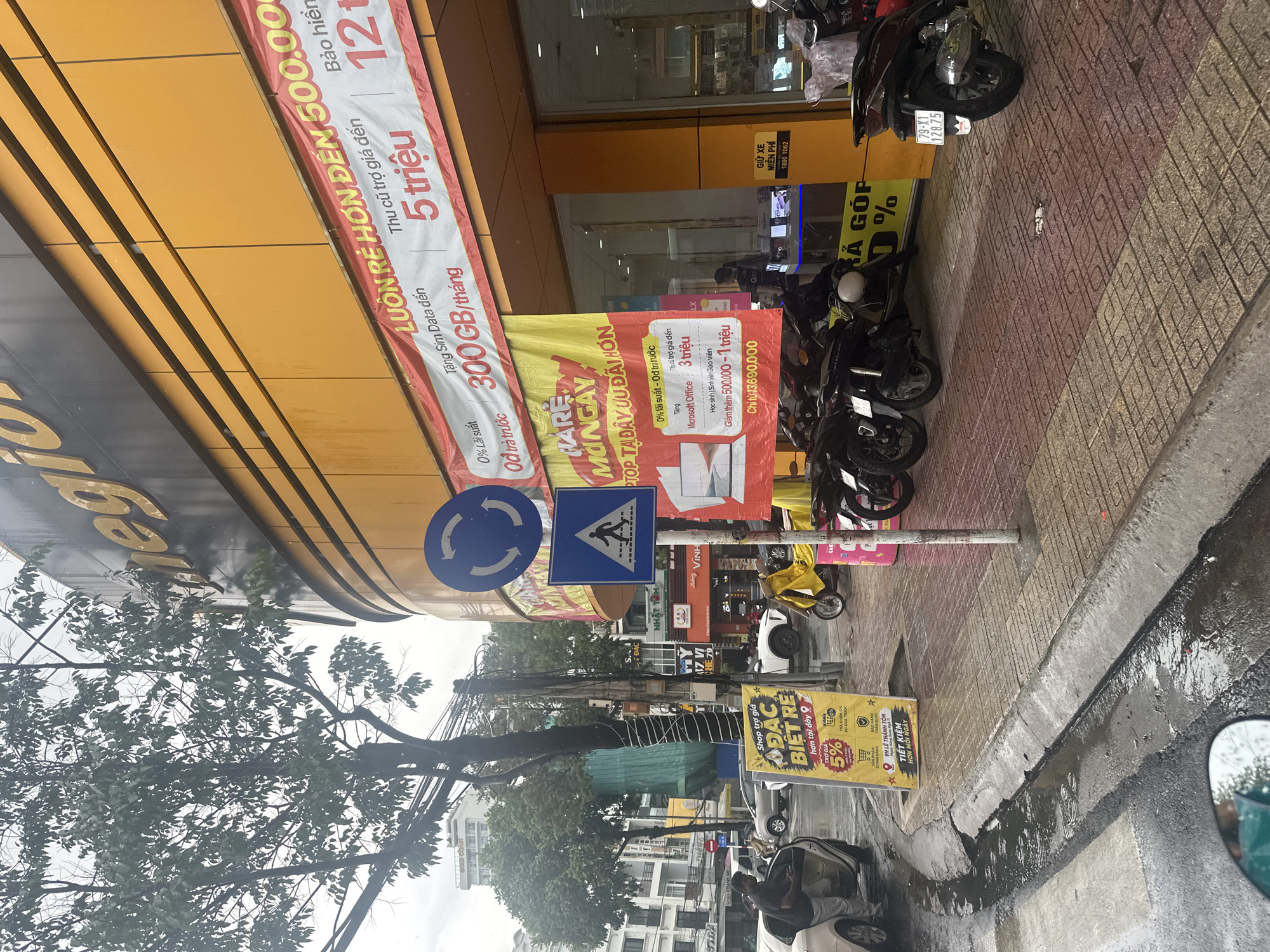 The image depicts a street scene in Nha Trang Ward, Vietnam, characterized by a mix of commercial activity and vehicular traffic. On the right side of the frame, motorbikes are parked along a tiled sidewalk adjacent to a street with water accumulating along the curb. A car is parked nearby with its door open and an individual is seen inside. Various signs and banners are prominently displayed along the storefront, advertising deals on mobile phones, data plans, and Microsoft Office. These signs include text in Vietnamese. A roundabout sign and a pedestrian crossing sign stand on a post near the sidewalk. Overhead, a tree branches out, intertwining with utility lines. The overall scene is urban and appears to be taken on a wet day.