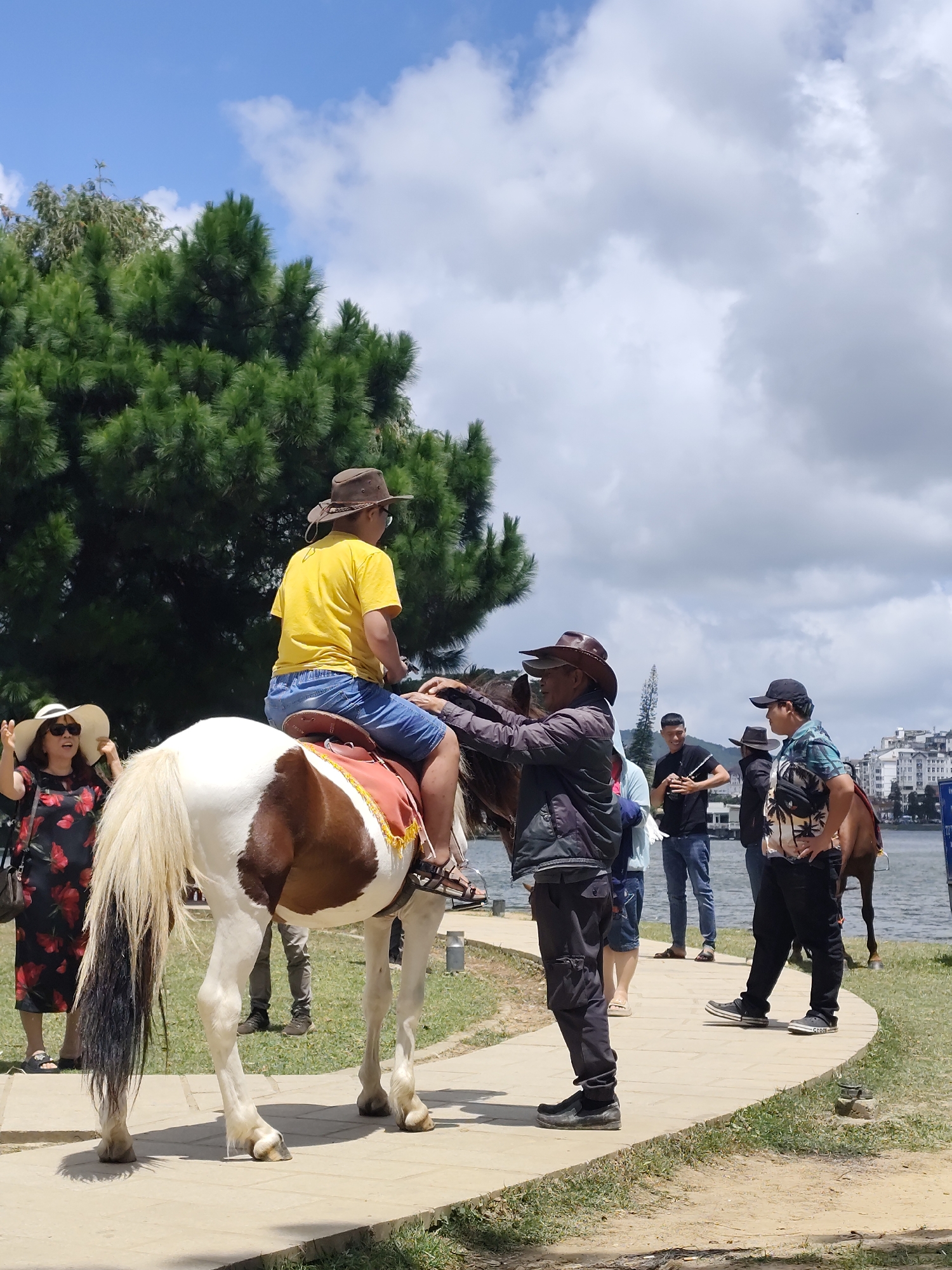 In Da Lat, Vietnam, on a paved pathway near a body of water, a group of people are gathered around a brown and white horse. A person wearing a yellow shirt and denim shorts, topped with a cowboy hat, is seated on the horse. Another individual, also in a cowboy hat, is standing next to the horse, appearing to assist the rider. To the left, a woman in a floral dress and a wide-brimmed hat gestures with her hands. Several other individuals are visible in the background, some standing near the water and others near buildings. Tall green trees form a backdrop against a sky with scattered clouds.