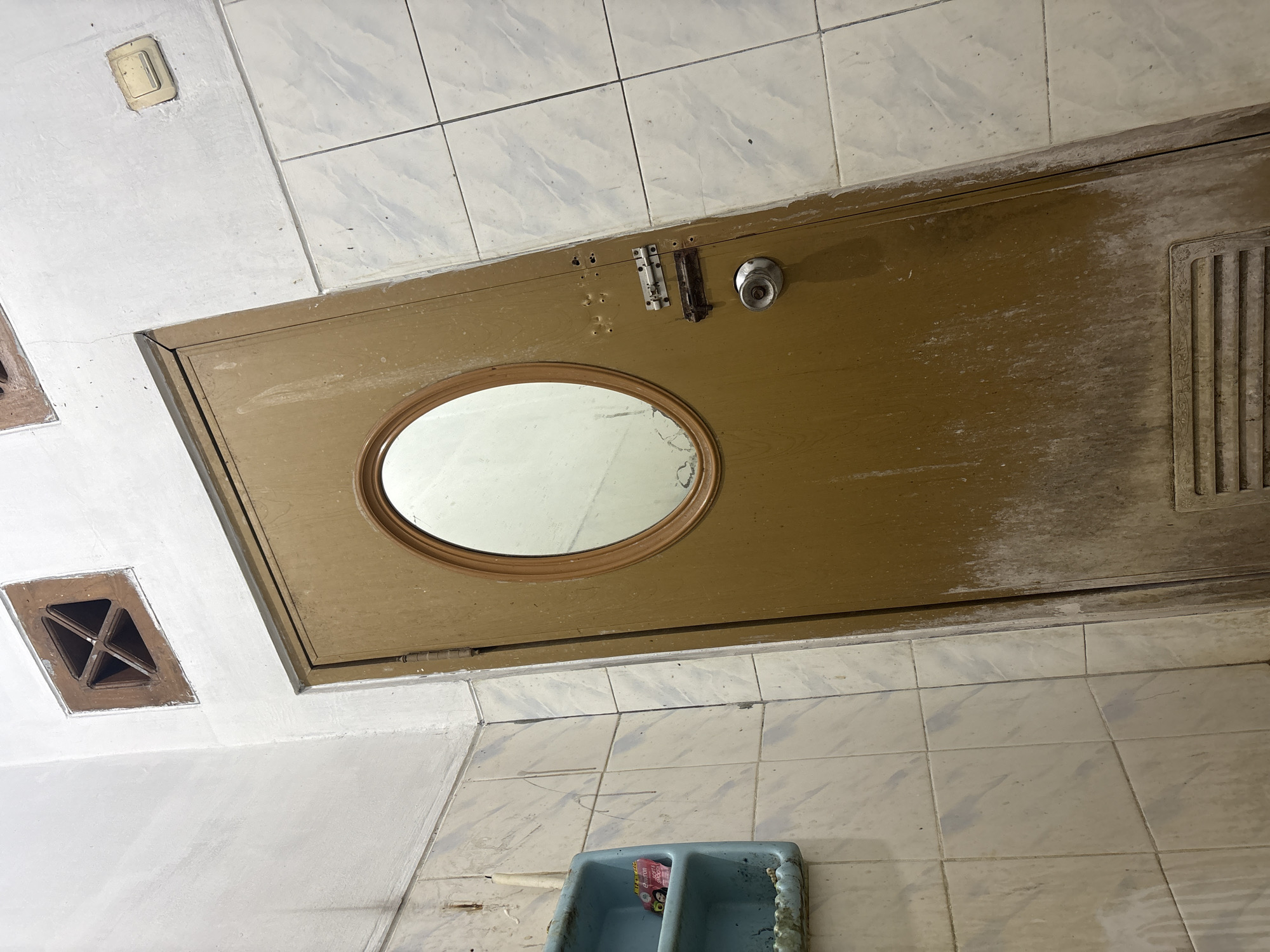 The image depicts a dingy interior, possibly a restroom, located in Palimanan, Indonesia. A light brown door with an oval-shaped mirror and a metal doorknob is prominently featured, tilted at an angle. The door is dirty, with visible streaks of grime and dust. The walls are tiled with a light-colored pattern, and some tiles appear to be stained. A blue plastic shelf with two small items is attached to the tiled wall on the left side of the door. Above the door, a square wooden vent with a cross pattern is visible in the white wall. To the right of the door, a light-colored electrical outlet is mounted on the wall. The overall scene suggests a neglected and possibly abandoned space.