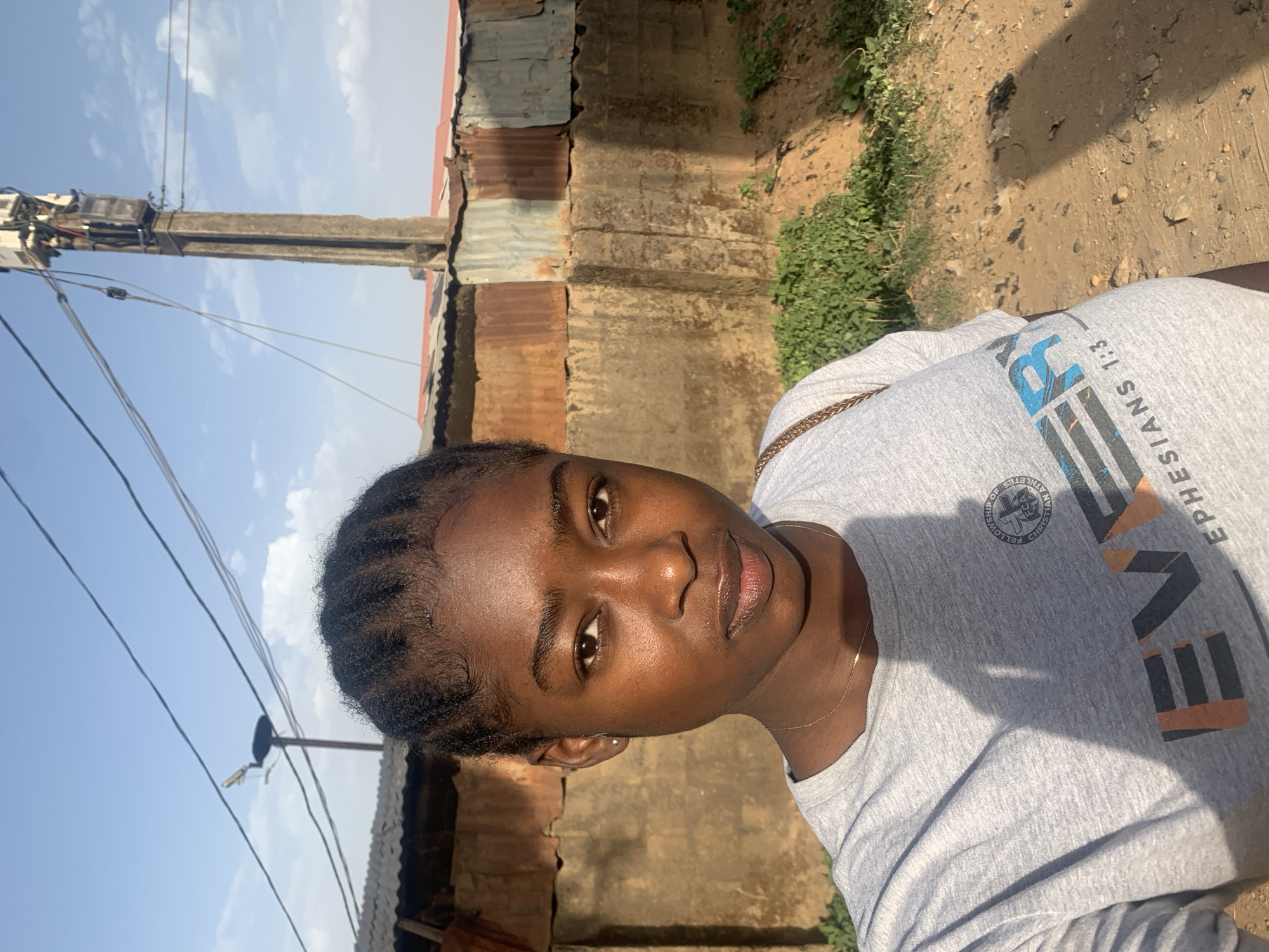 A medium, outdoor shot depicts a young woman in Rafin Par, Nigeria. She is positioned slightly to the right of the frame, facing the camera. She has dark skin, dark hair styled close to her head, and wears a light gray t-shirt with lettering. A thin gold necklace is visible around her neck.
The background includes a weathered building with a combination of concrete and corrugated metal. A utility pole with wires extends into the sky on the left side of the frame. Ground covered with dirt and some sparse vegetation is seen in the lower background. The sky is partially visible with scattered clouds. The image has a bright, natural light.