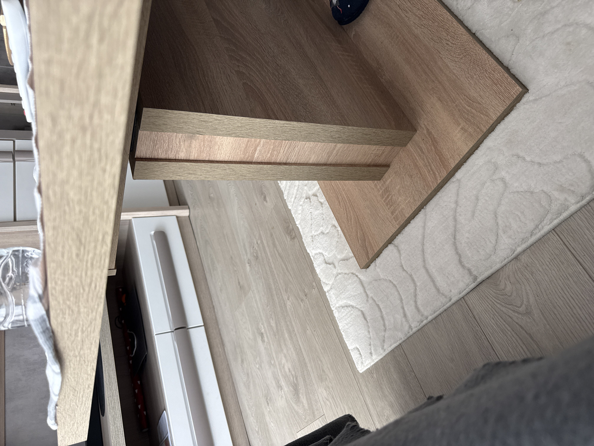 This image shows a close-up view of a room interior. A wooden desk or table is partially visible, with its edge resting on a white, plush rug. The rug is on top of a light-colored wooden floor. Part of a white cabinet with drawers is visible to the left. The scene appears to be indoors, possibly in a home. It is impossible to tell if this is in Lovech, Bulgaria. The image lacks any identifiable people or actions.