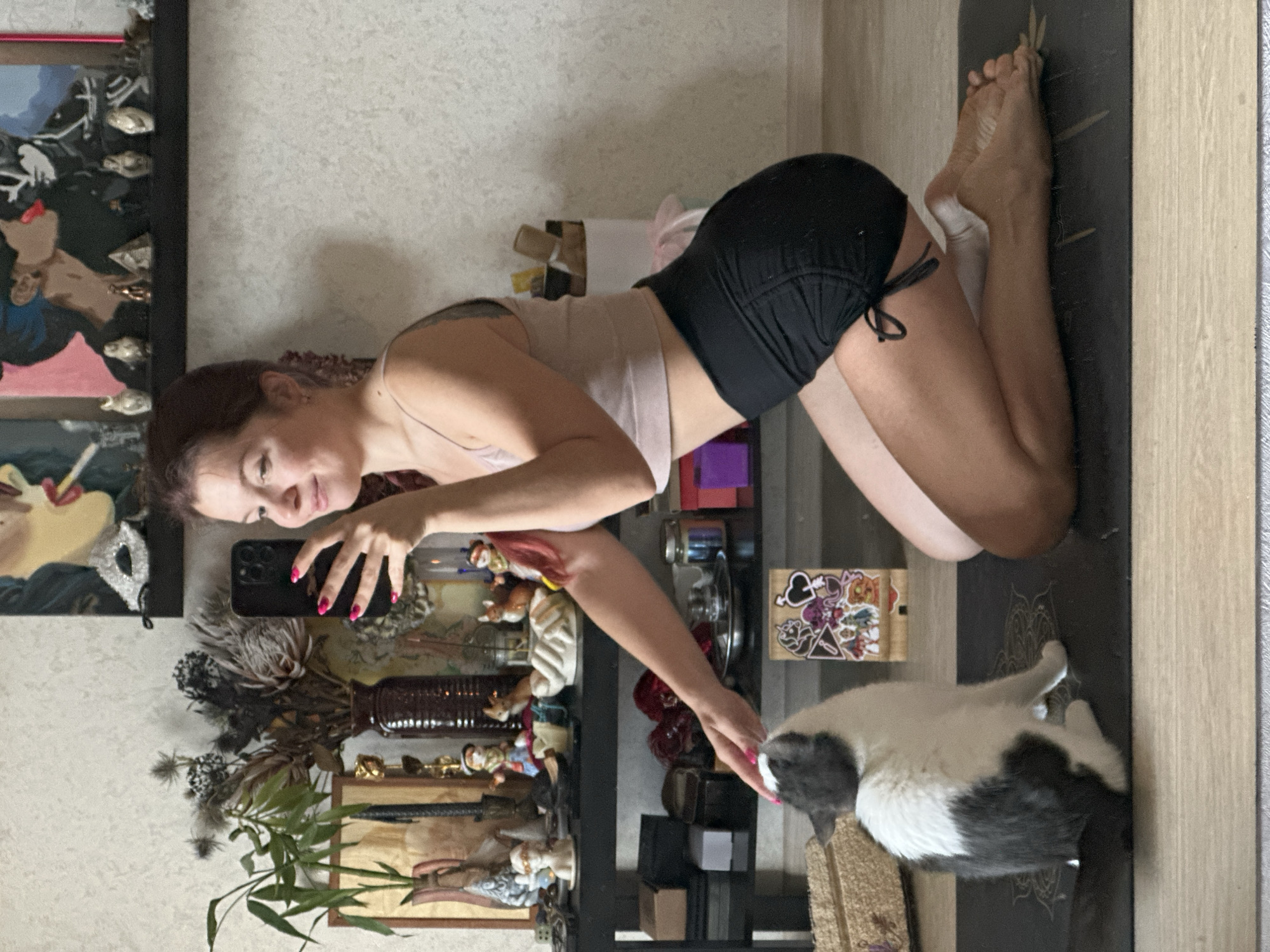 A woman is kneeling on a yoga mat, taking a selfie with her phone. She has her left hand extended, petting a grey and white cat. The cat is sitting on the yoga mat, looking up at the woman. The woman is wearing a light pink tank top and black shorts. She has red nail polish on her fingers. In the background, there are shelves filled with various decorative items, including vases, figurines, and artwork. The wall behind the shelves is textured. The floor appears to be light-colored wood. The scene takes place indoors, likely in a residential setting. There is no information to suggest this scene is located in the city of Vladimir, Russia.