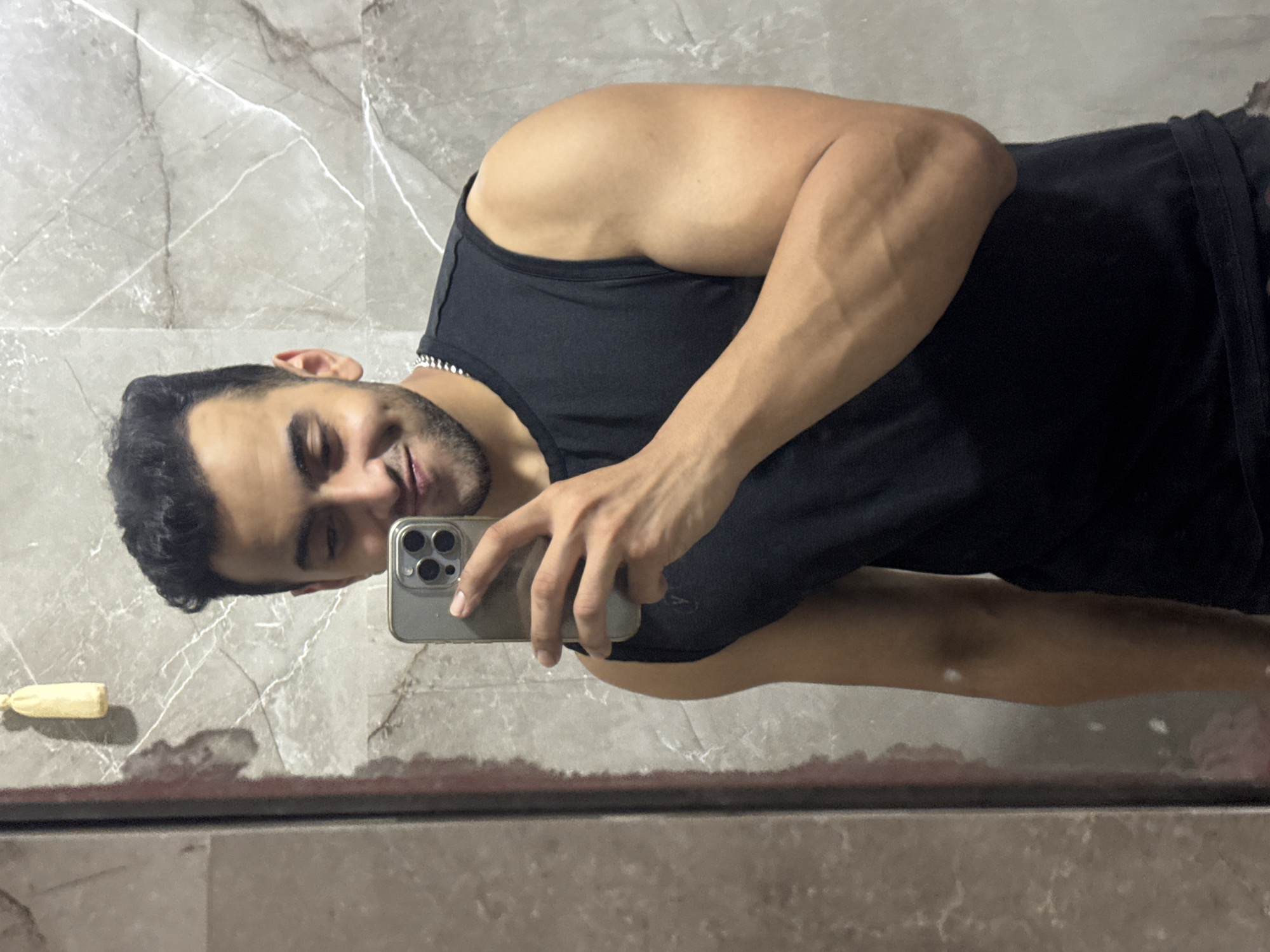 A man is taking a selfie in a bathroom with gray marble walls. He is wearing a black tank top and holding an iPhone with three lenses in his right hand. There is a small, pale yellow object to his left, near the edge of the mirror or reflective surface. The man has dark hair, a light beard, and appears to be in his late 20s or early 30s. The location is not discernible from the image.