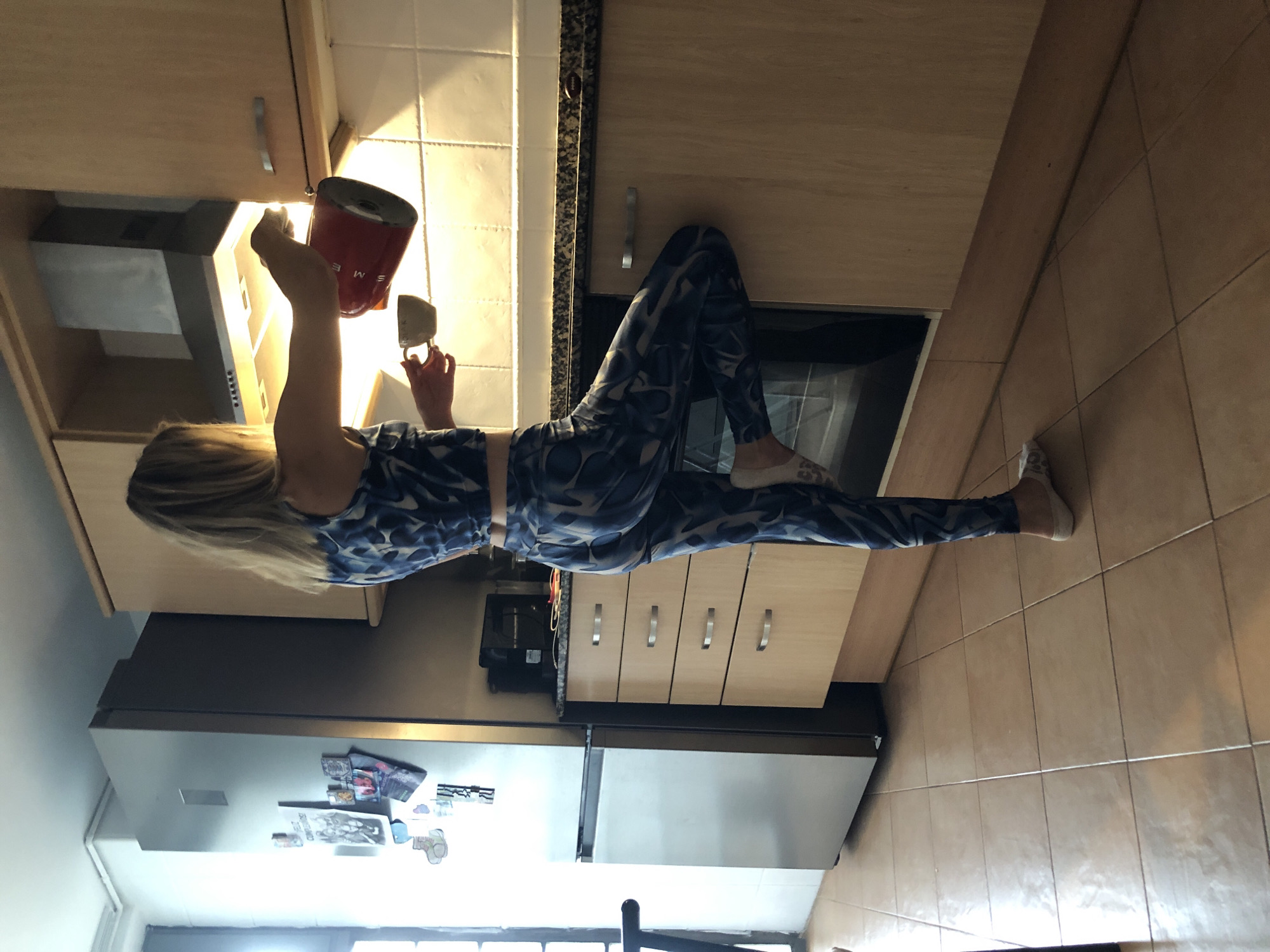 A woman with blonde hair, wearing a blue and white patterned top and matching leggings, is in a kitchen. She is pouring liquid from a red kettle into a white cup, which she holds in her left hand. The kitchen has light wood cabinets, a granite countertop, and tiled floors. A silver refrigerator is visible to the left of the woman, and a range hood is above the counter. The scene appears to be an indoor residential kitchen in the city of València, Spain.