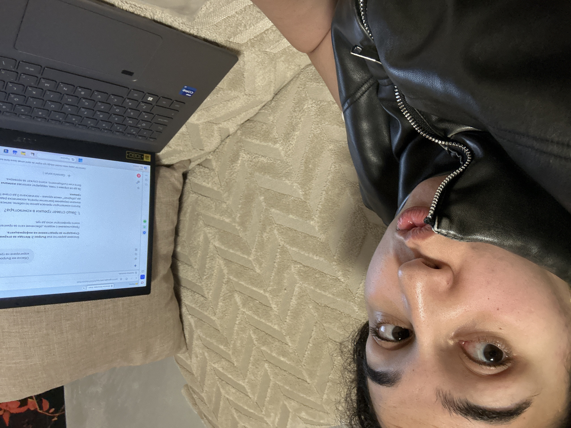 Here is a detailed description of the image:
**Scene:** The image appears to be a selfie taken from a slightly above perspective. A person is lying on a light-colored, patterned cushion or bed. To the left of the person's face is a laptop.
**People:** One person is visible, with their face being the primary focus. They have dark hair, dark eyebrows, and light skin. They seem to be wearing a black leather jacket with a zipper near their mouth.
**Objects:** The main objects include:
* A laptop: A grey laptop with a screen displaying text. It's partially open, revealing the keyboard.
* A cushion/bed: The person is lying on a textured, light-colored surface.
* A black leather jacket.
**Actions:** The person is lying down and seems to be either taking a picture of themselves or has set up a camera. The person is in front of the laptop.
**Interactions:** The person is interacting with or looking at the laptop.
**Environment:** The environment seems to be indoors, possibly at the person's home.
There is no indication in the image where this photo was taken, so I cannot determine the location in Burgas, Bulgaria.