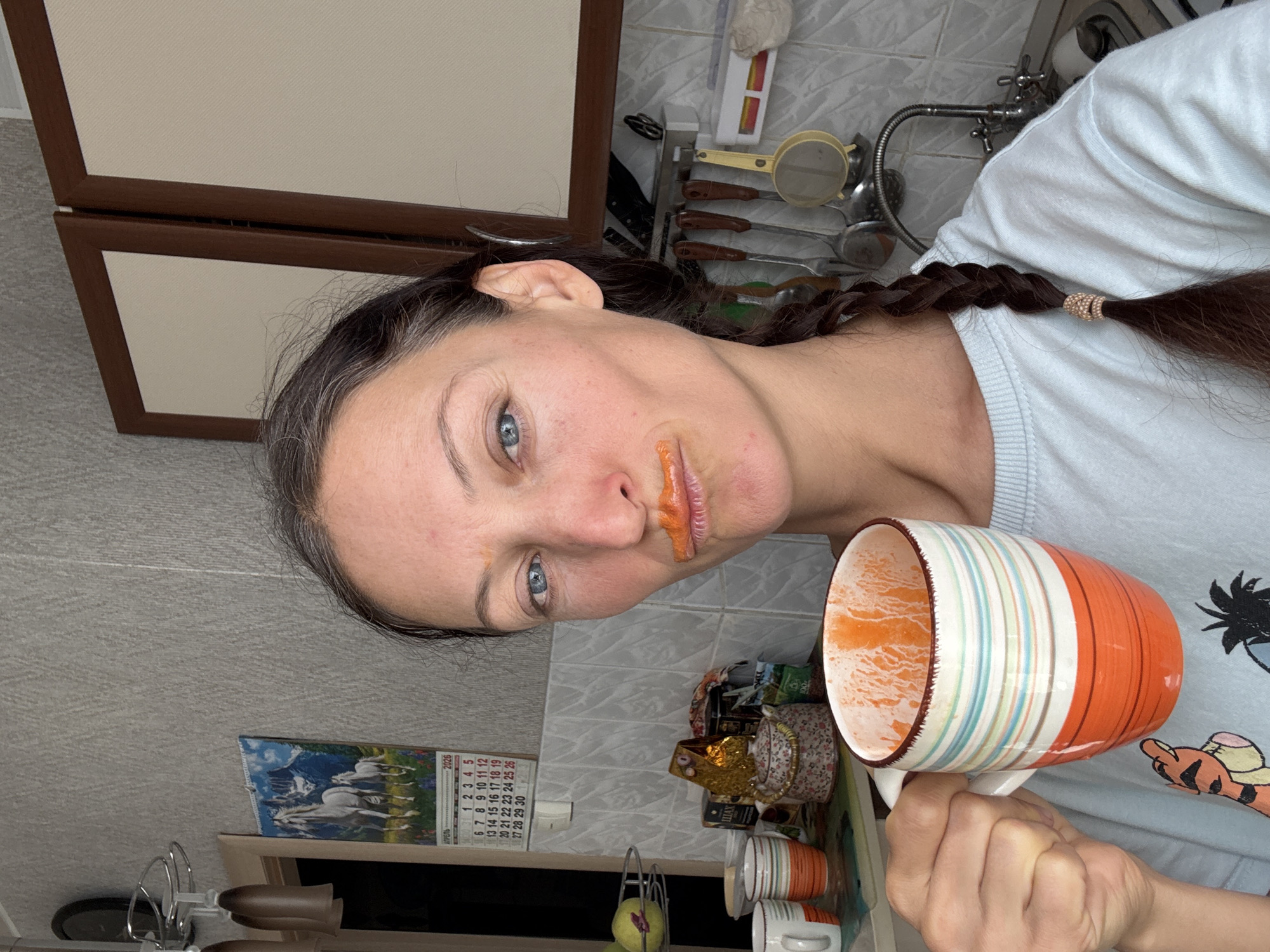 A casual portrait captured in a domestic kitchen, likely in Ulyanovsk, Russia, shows a woman with dark hair tied in a braid and clear blue eyes. The image is tilted, presenting her face slightly upward towards the left. A notable detail is the vivid orange substance smudged around her mouth, indicating she has recently consumed a beverage or food of that color.
In her right hand, she holds a striped mug, adorned with shades of orange, white, and light blue/green, which also contains an orange residue. She is wearing a light blue t-shirt with a partial graphic of what appears to be a cartoon character (possibly Tigger) on the chest.
The background reveals a typical kitchen environment with light-colored tiled walls, a sink, and a faucet on the right side. Various kitchen utensils, including spoons, a sieve, and a knife block, hang neatly above the counter. Dark-framed cabinets with light panels are visible in the upper left. On the counter, several other cups, possibly tea boxes, and a decorative ceramic item are arranged.
A visible calendar on the wall displays "АПРЕЛЬ" (April) and "2026," with the dates 1 through 30 clearly marked. The calendar's image features white horses in a mountainous landscape. The overall scene depicts a relaxed, everyday moment, possibly during a break or after a meal. The woman's expression is neutral, conveying a sense of casualness.