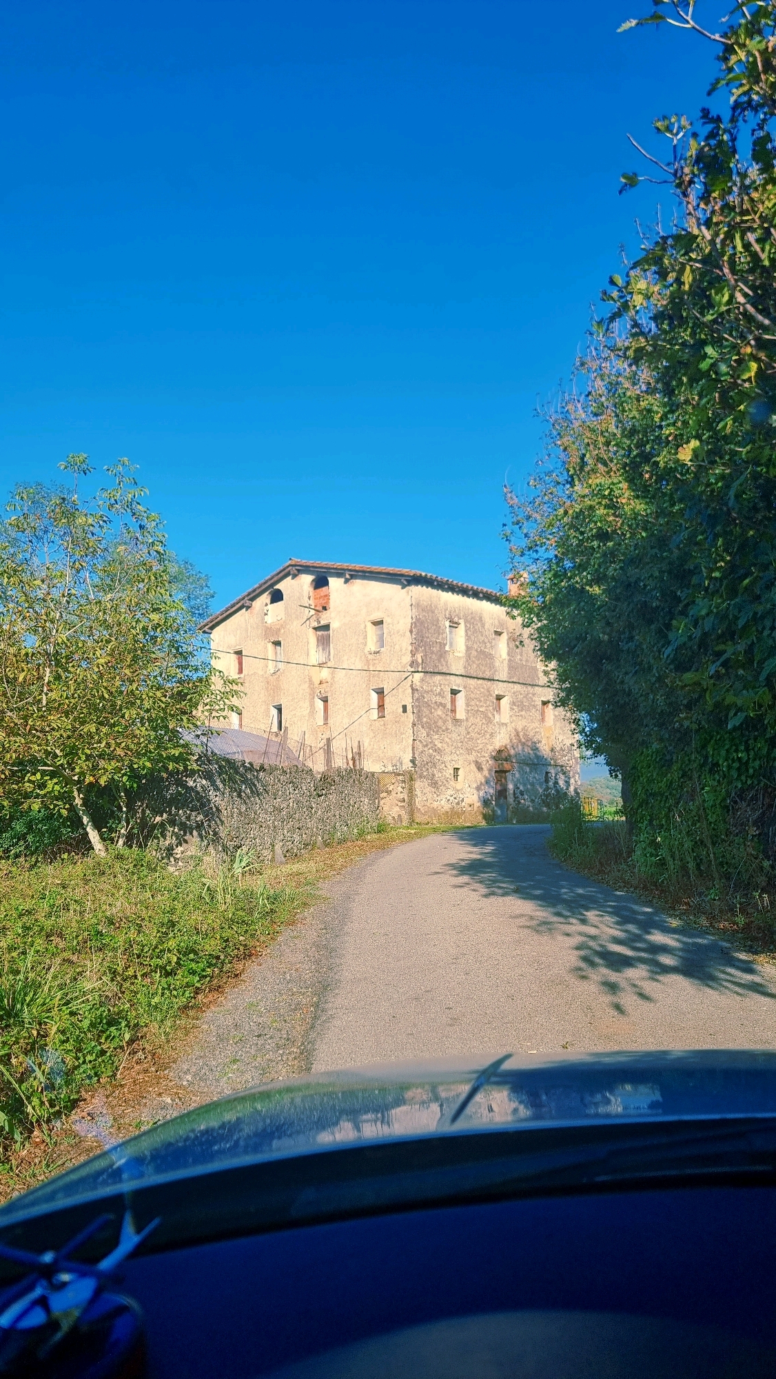 The image captures a rural scene in Sant Joan les Fonts, Spain, featuring a weathered, two-story stone building situated alongside a narrow asphalt road. The building, appearing to be an old farmhouse or manor, has numerous rectangular windows, some of which are dark or appear boarded up. There are also several arched openings on the upper level, one of which shows exposed brickwork. A stone wall runs along the side of the road in front of the building, with vegetation growing on it and along the roadside. Lush green trees and foliage line the road, casting shadows from the sunlight. The sky is a clear, vibrant blue, indicating a sunny day. The foreground of the image is occupied by the hood and windshield of a dark-colored vehicle, suggesting the photo was taken from inside or very close to a car. No people are visible in the scene.