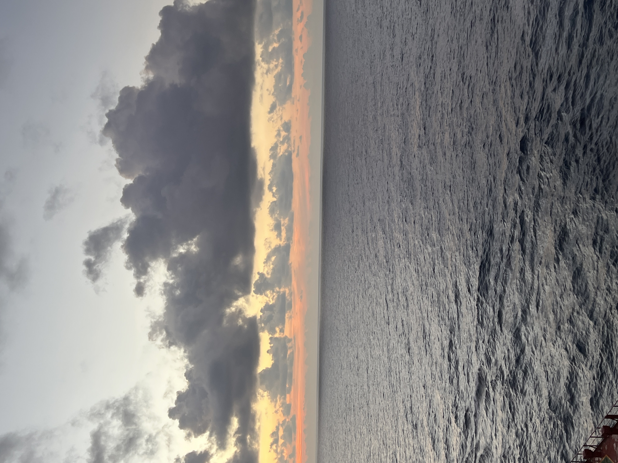 The image shows a body of water under a sky with clouds. The water is dark with small ripples. The sky transitions from a pale gray at the top to hues of orange and yellow near the horizon, where clouds are present. A small portion of a red and white structure, possibly part of a ship or dock, is visible in the bottom left corner. The location is Unknown, Unknown.