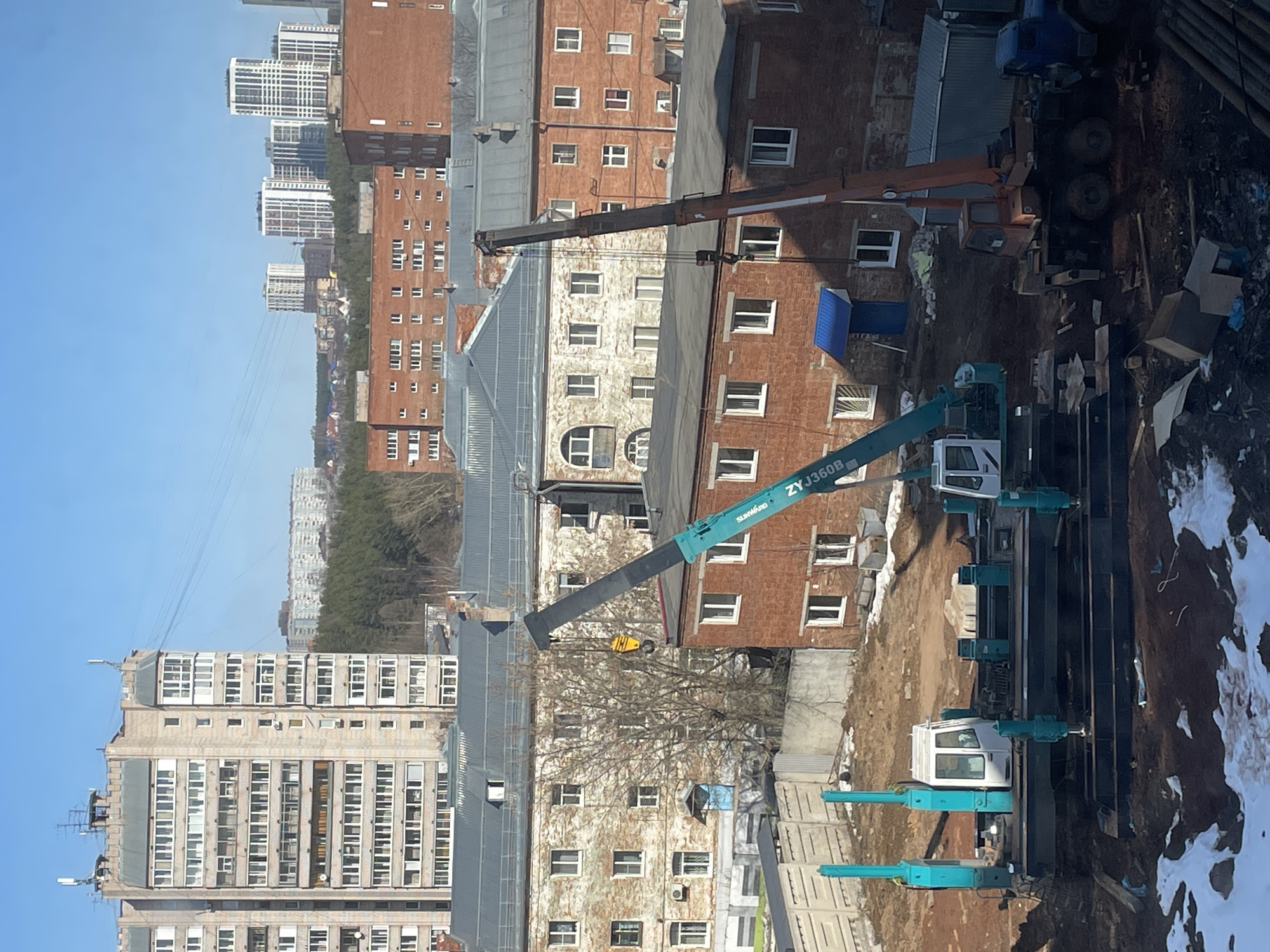 The image shows an urban construction site in Izhevsk, Russia. The scene includes construction equipment, specifically two cranes, positioned amidst several multi-story brick and concrete buildings. One crane is blue and white with the label "ZYJ360B." The second crane is brown and on a flat bed truck. Large metal beams and construction materials are present on the site. Patches of snow are visible along the edge of the construction area. Tall buildings and trees are visible in the background. The sky is clear blue.