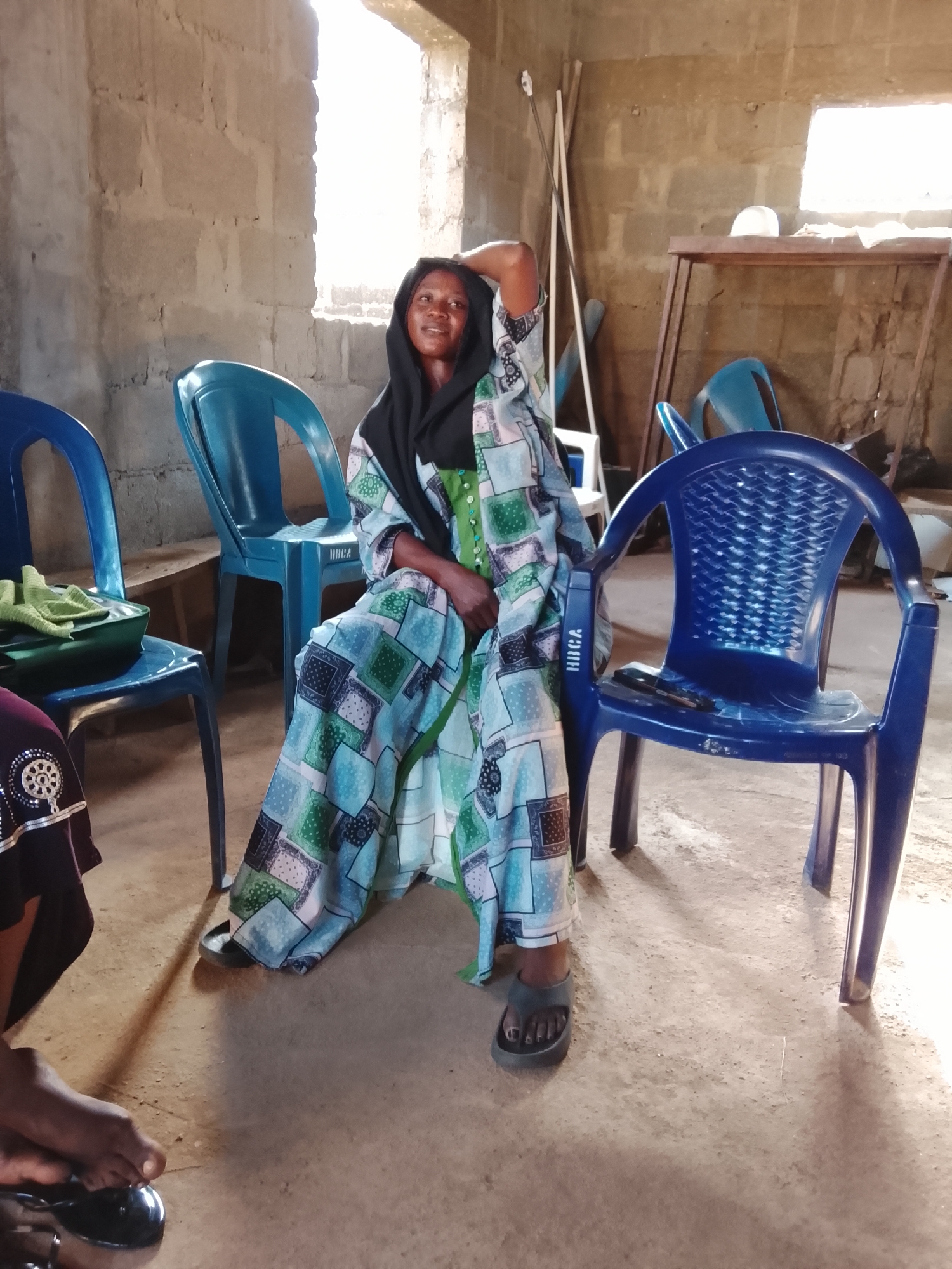 The image depicts a woman in Akwanga, Nigeria, sitting on a blue plastic chair. She is wearing a patterned dress, a green undershirt, and a black head covering. Her left arm is raised behind her head. The setting appears to be an indoor space with concrete block walls. Several other blue plastic chairs are visible, along with a metal structure in the background. The floor is a light-colored concrete.
