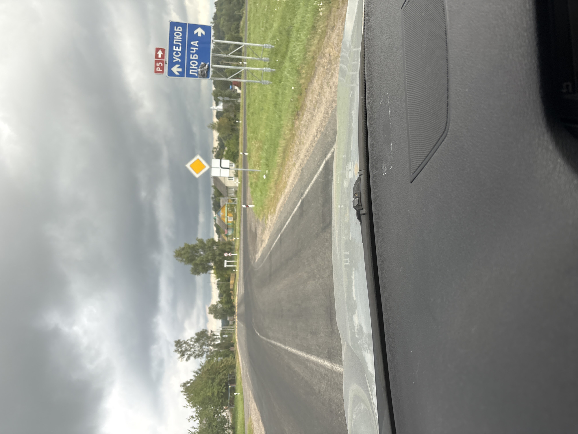 The image shows a view from inside a vehicle, looking out at a rural road intersection in the direction of Уселюбскі сельскі Савет, Belarus. The sky is overcast with dark clouds. On the right side of the road, there are blue directional signs indicating "УСЕЛОБ" (USELOV) with an upward arrow and "ЛЮБЧА" (LYUBCHA) with a rightward arrow. Above these signs, there is a smaller red sign with "P5" and a rightward arrow. A yellow diamond-shaped sign is also visible on the left side of the road. The road itself appears to be paved and has white lane markings. In the background, there are trees, houses with yellow and beige walls, and a white church spire. The foreground of the image is dominated by the interior of the car, specifically the dashboard. There are no people or animals visible in the scene.