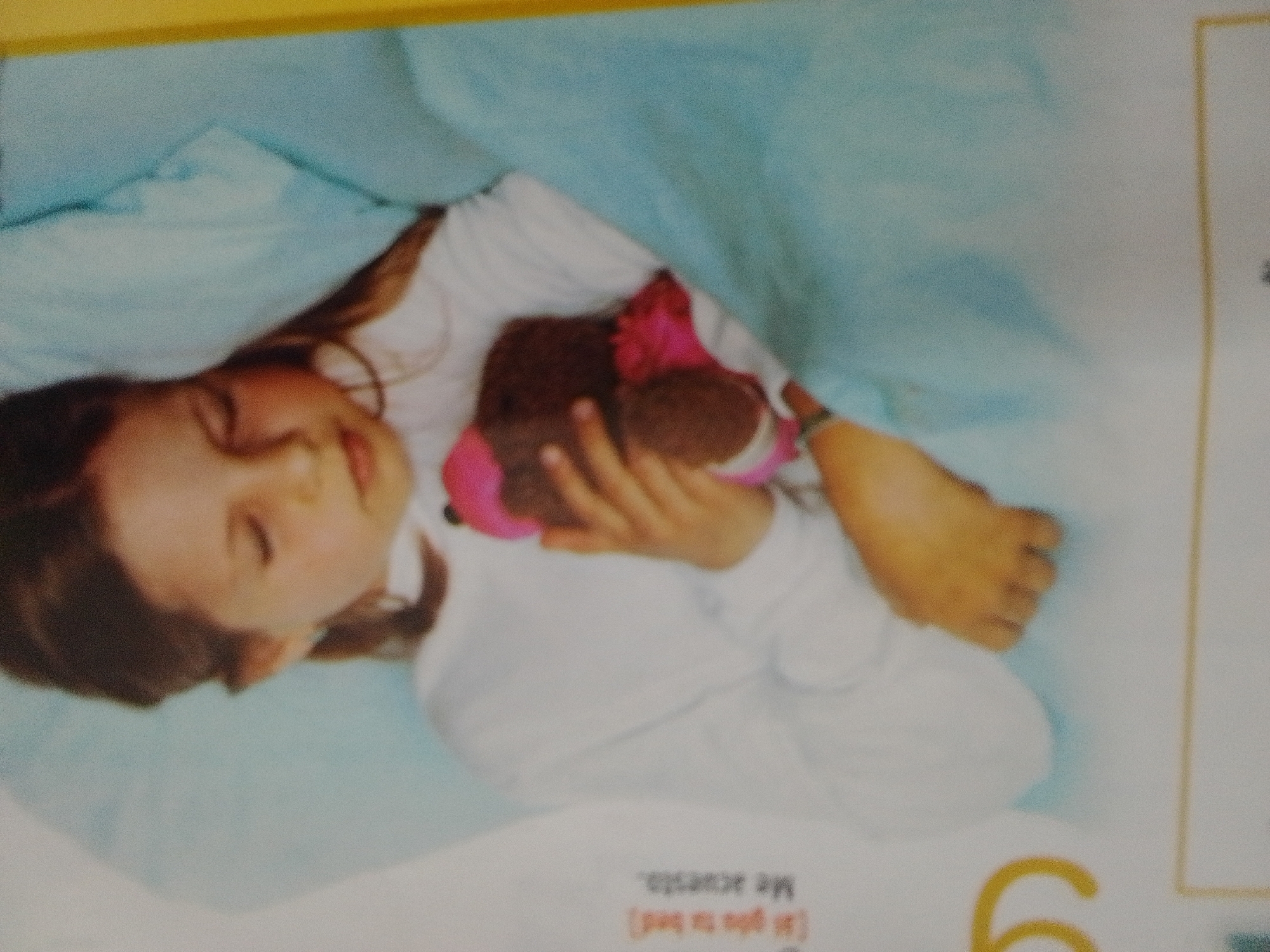 The image shows a child sleeping under a light blue blanket, holding a stuffed animal. The child is on their side, with their face turned away from the camera, and their eyes are closed. The stuffed animal is brown and pink. The background is a plain white surface. The image appears to be a photograph of a child in bed. There is no information in the image to suggest a location in La Tagua, Venezuela.