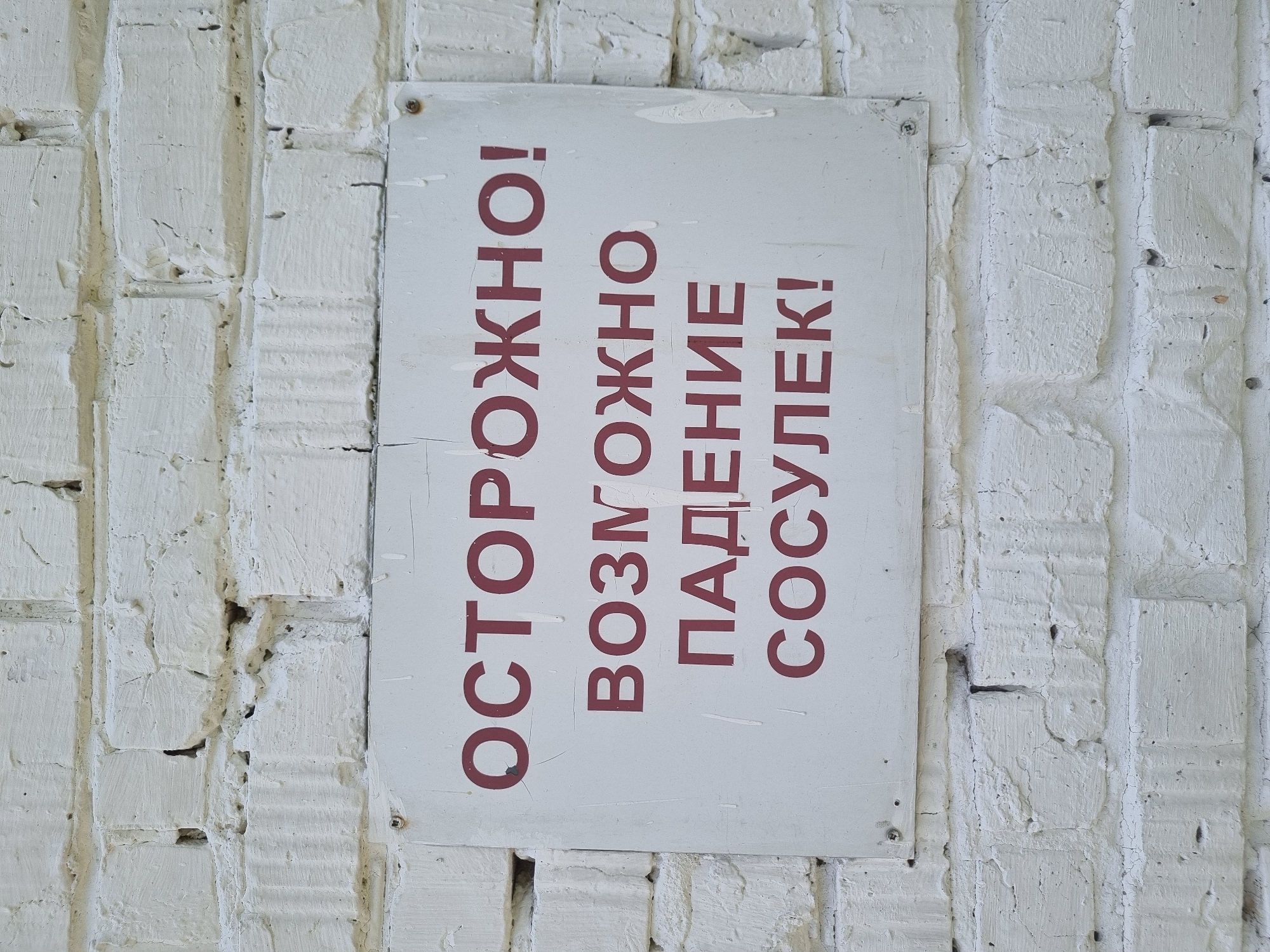 A close-up shot shows a white brick wall with a warning sign in the city of Kemul, Russia. The sign, attached to the wall with screws, is white with red lettering in Russian, which translates to "Caution! Possible icicle fall!". The wall appears aged, with chipped paint and visible grout lines between the bricks. The scene depicts a potential winter hazard in an urban environment.