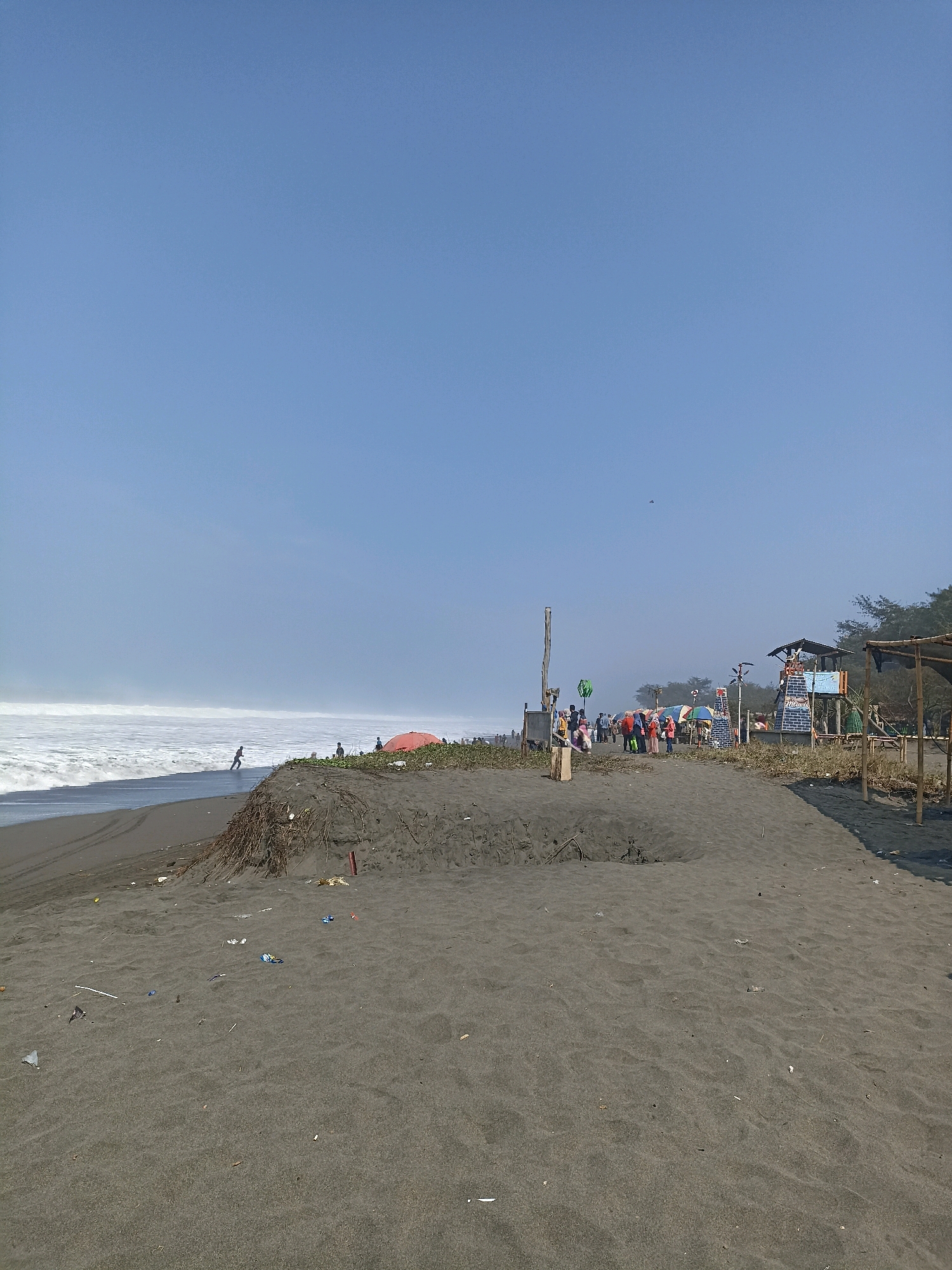 This is a medium-shot, eye-level, outdoor photograph taken on a sunny day at a beach in Kenoyajaya, Indonesia. The beach features dark sand and is lined with vegetation and some man-made structures. In the foreground, the sand is rippled and shows signs of litter. There is a large mound of sand and debris to the left. In the midground, there are several people, some standing in small groups, and others gathered around what appear to be vendor stalls and an elevated lifeguard tower. A red umbrella is visible amongst the crowd. The ocean is in the background, with large waves breaking on the shore. A lone figure is seen running into the water on the left side of the frame. The sky is clear and blue.