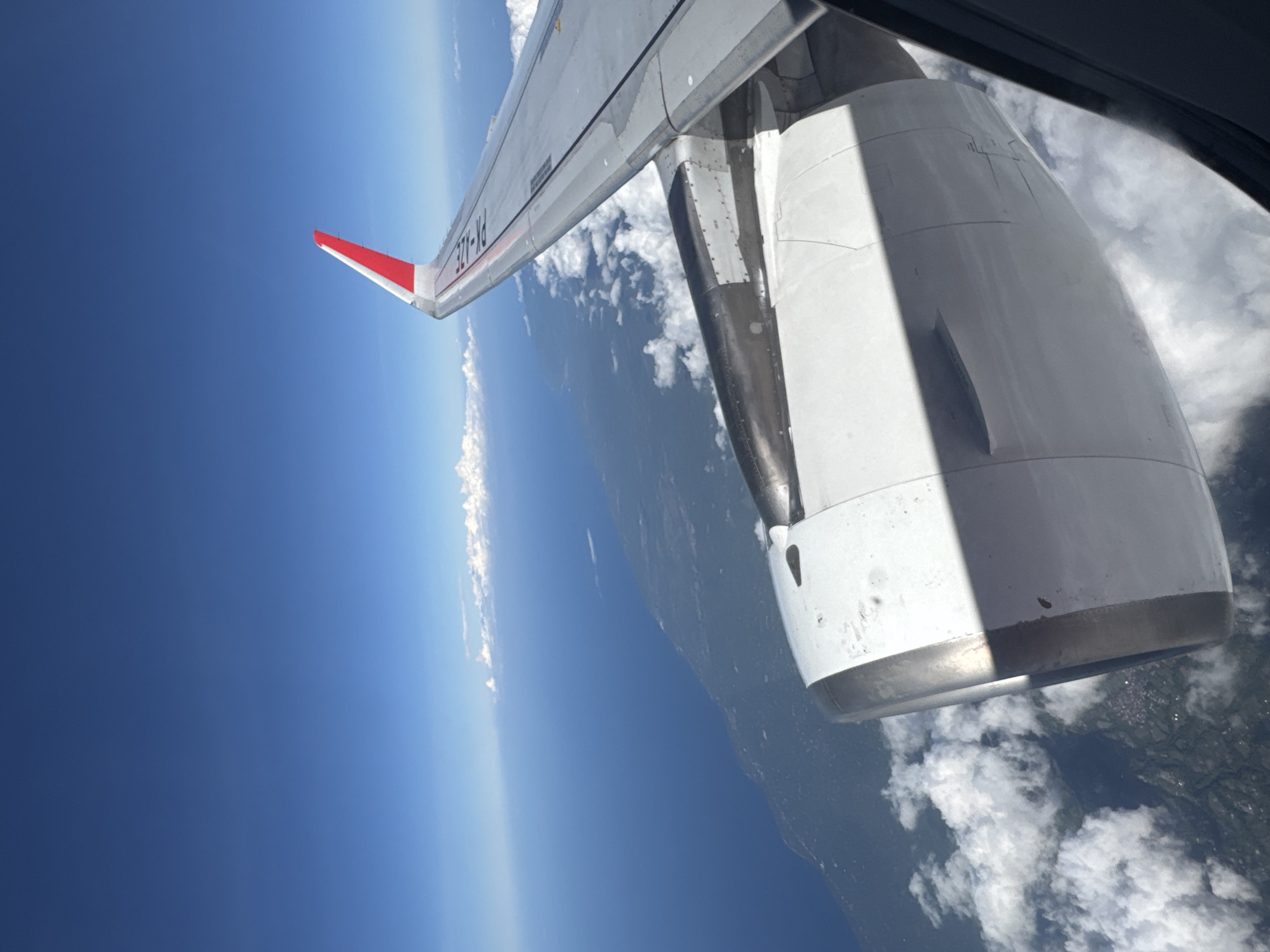 A view from an airplane window shows the wing and engine over a landscape of clouds, forest, and a clear blue sky. The wing has a red and white tip. The engine is white with some gray and black components. The background landscape reveals a horizon and some visible land. The plane is flying over Bangli, Indonesia.