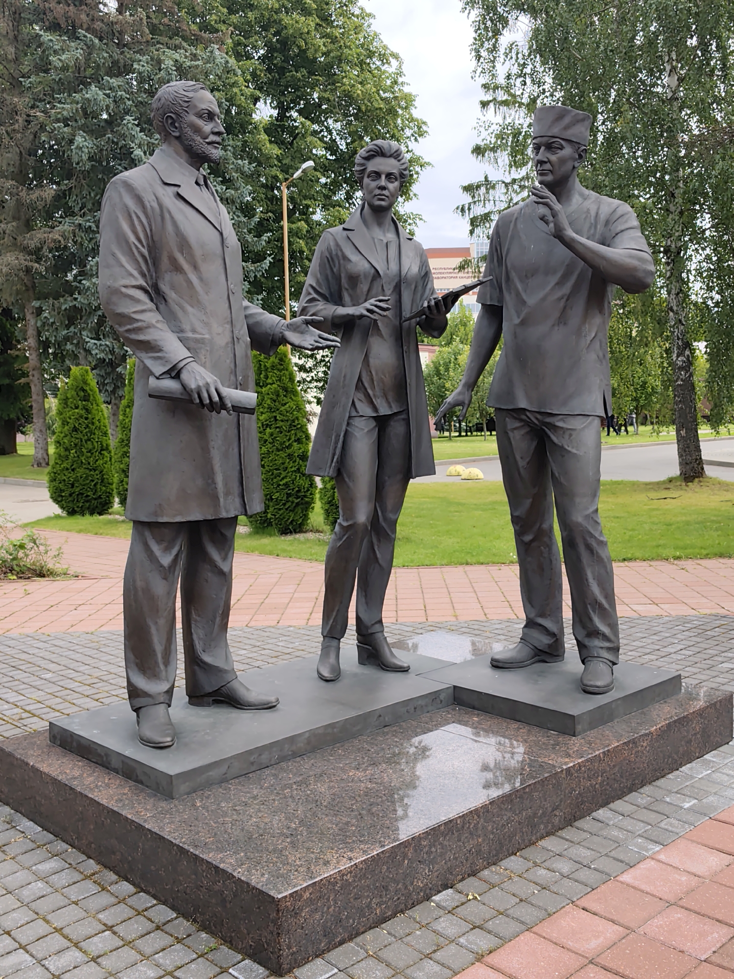 A bronze sculpture of three medical professionals stands on a stone pedestal in a park-like setting. The sculpture features a man on the left wearing a long coat and holding a rolled-up document. In the center is a woman in medical attire, holding a tablet. To the right, a man in medical scrubs and a cap is gesturing with his hand. The figures appear to be engaged in a discussion or presentation. The sculpture is situated on a multi-tiered base, with the bottom layer made of speckled dark granite and the upper layers of smooth, dark stone. The surrounding area is paved with interlocking bricks and features a grassy lawn with scattered trees and manicured bushes. In the background, a building with visible text, identified as "РЭСПУБЛІКАНСКІ НАВУКОВЫ ПРАКТЫЧНЫ ЦЭНТР АНЭСТЭЗІЯЛАГІІ І ІНТЭНСІЎНАЙ ТЭРАПІІ" (Republican Scientific and Practical Center of Anesthesiology and Intensive Care), is visible, confirming the location in the Baravliany rural council, Belarus. The sky is overcast.