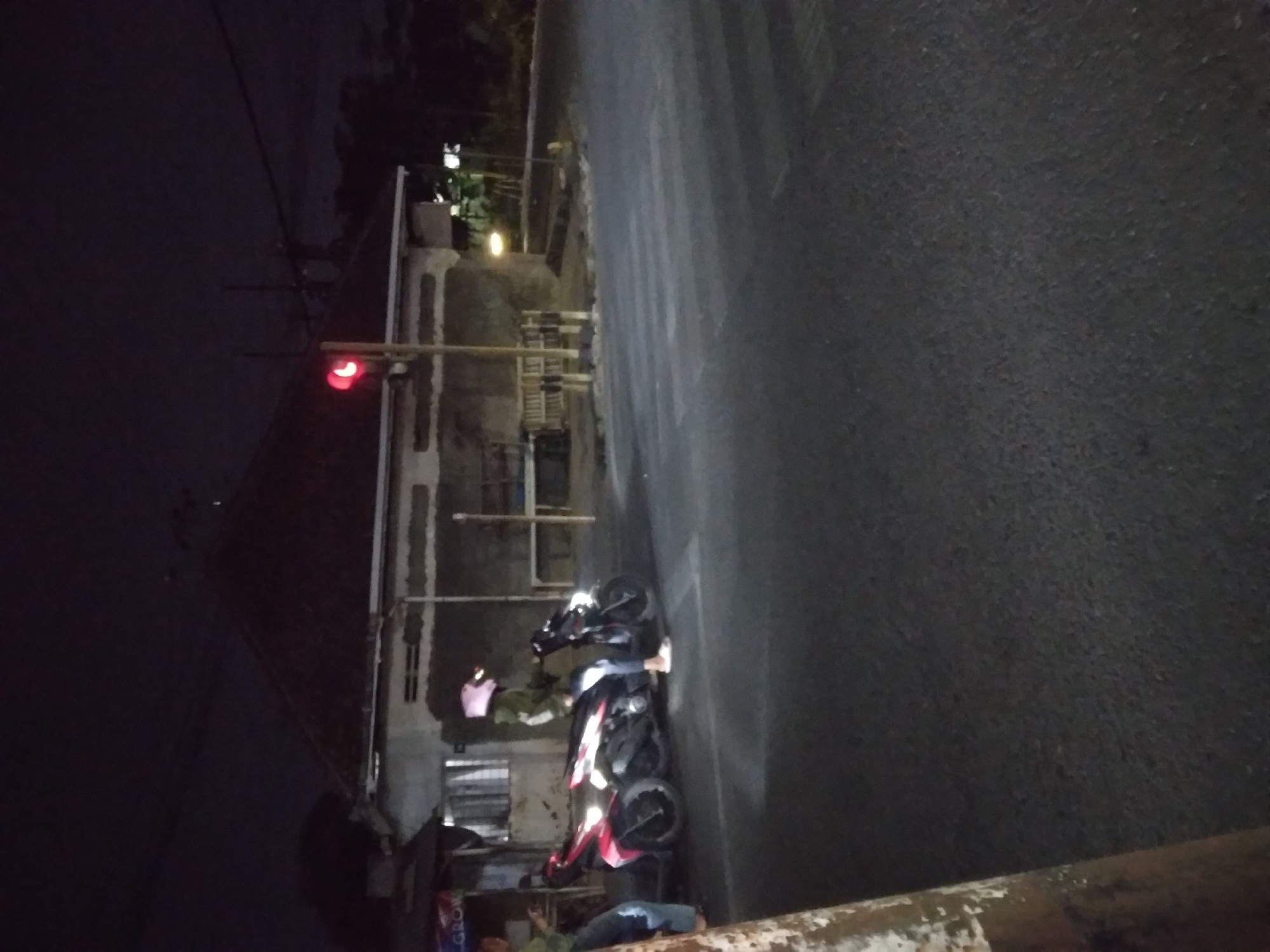 The image depicts a night scene at an intersection in Mersi, Indonesia. A traffic light is illuminated red. Two motorcycles are parked at the intersection. One motorcycle is pink and the other is black. A person wearing a pink helmet and a green jacket is sitting on the black motorcycle. Another person is partially visible on the left side of the image, standing next to the pink motorcycle. A building with a dark roof is visible in the background. The ground is paved, and a crosswalk is marked on the road. The overall environment appears to be a street intersection with some commercial structures.