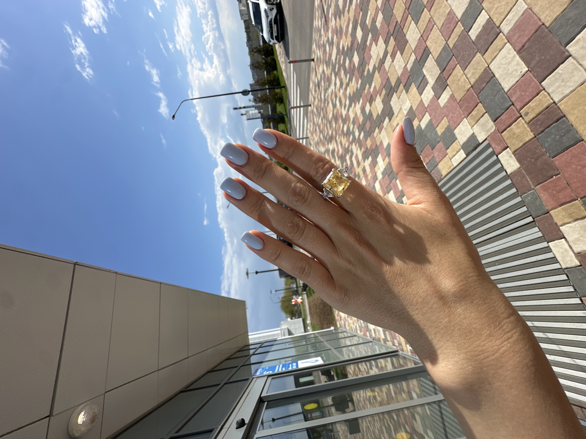 The image shows a person's left hand wearing a ring with a large yellow stone and smaller clear stones. The hand is positioned in front of a building with a gray exterior and glass windows. The person has light blue nail polish. The background includes a blue sky with scattered clouds and a multicolored brick pathway. A street and cars are visible in the distance. The location appears to be an urban environment, but the specific city is unknown.