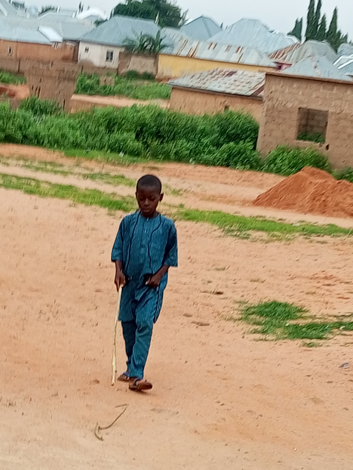 A young boy, wearing a blue traditional outfit, walks across a dusty, unpaved road. He holds a long stick in his right hand and appears to be using it as a cane. The boy is looking down at the ground as he walks. Behind him, a pile of reddish-brown dirt is visible, along with sparse green grass and several buildings under construction. The buildings have brick walls and corrugated metal roofs. The sky is overcast. The image was taken in Tumfure, Nigeria.