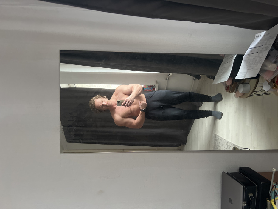 An adult male with curly blonde hair and a mustache is captured in a full-length mirror selfie within an indoor residential setting in Saint Petersburg, Russia. The man, exhibiting a muscular physique, stands shirtless, wearing black sweatpants and grey socks. He holds a silver smartphone at waist height with both hands, angled to photograph his reflection while flexing his biceps. A dark smartwatch is visible on his left wrist.
The mirror's reflection shows a room with dark grey curtains drawn across what appears to be a window on the right. A white radiator pipe is visible along the lower wall, and a small, light-colored shelf containing bottles or jars is affixed to the wall to the right of the man. The floor is light-colored, partly covered by a small, light-patterned rug.
Outside the mirror's frame, on the left, a silver laptop rests atop a black case. On the right, a small, dark, round side table holds scattered white papers, small containers, and other miscellaneous items. The surrounding walls are painted an off-white or light grey.