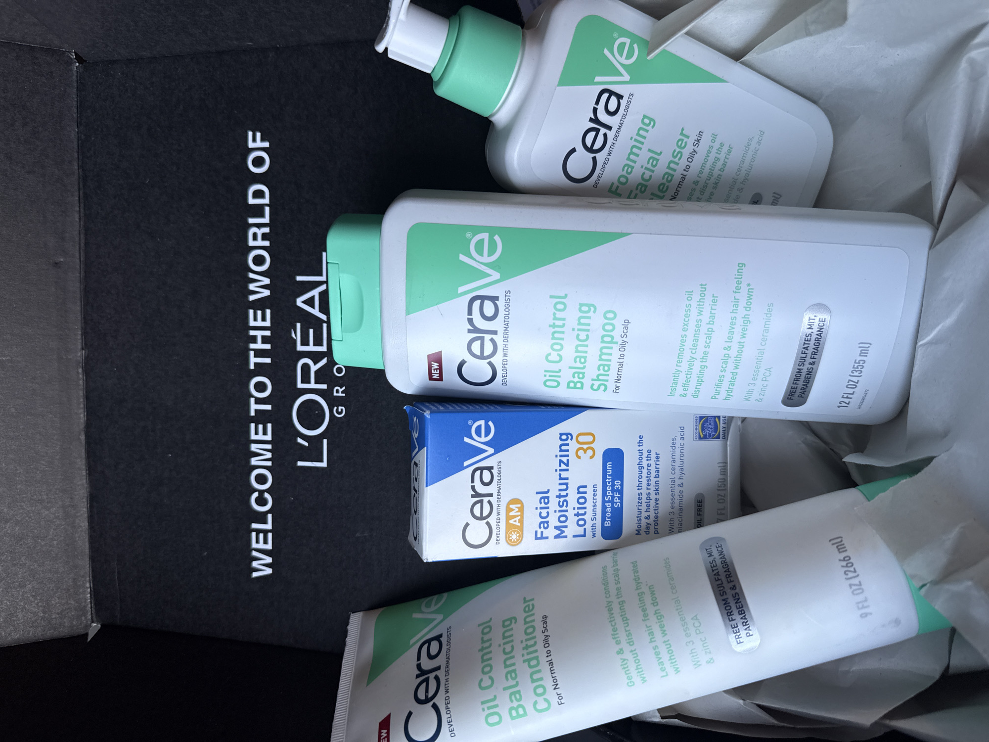 The image shows a collection of Cerave skincare products, including a foaming facial cleanser, oil control balancing shampoo, oil control balancing conditioner, and facial moisturizing lotion, arranged inside a black box labeled "WELCOME TO THE WORLD OF L'ORÉAL GROUP". The products are positioned at various angles within the box. The scene appears to be indoors, likely a home or storage area. The location cannot be determined from the image, and thus I cannot say if this image was taken in Columbus, United States of America.