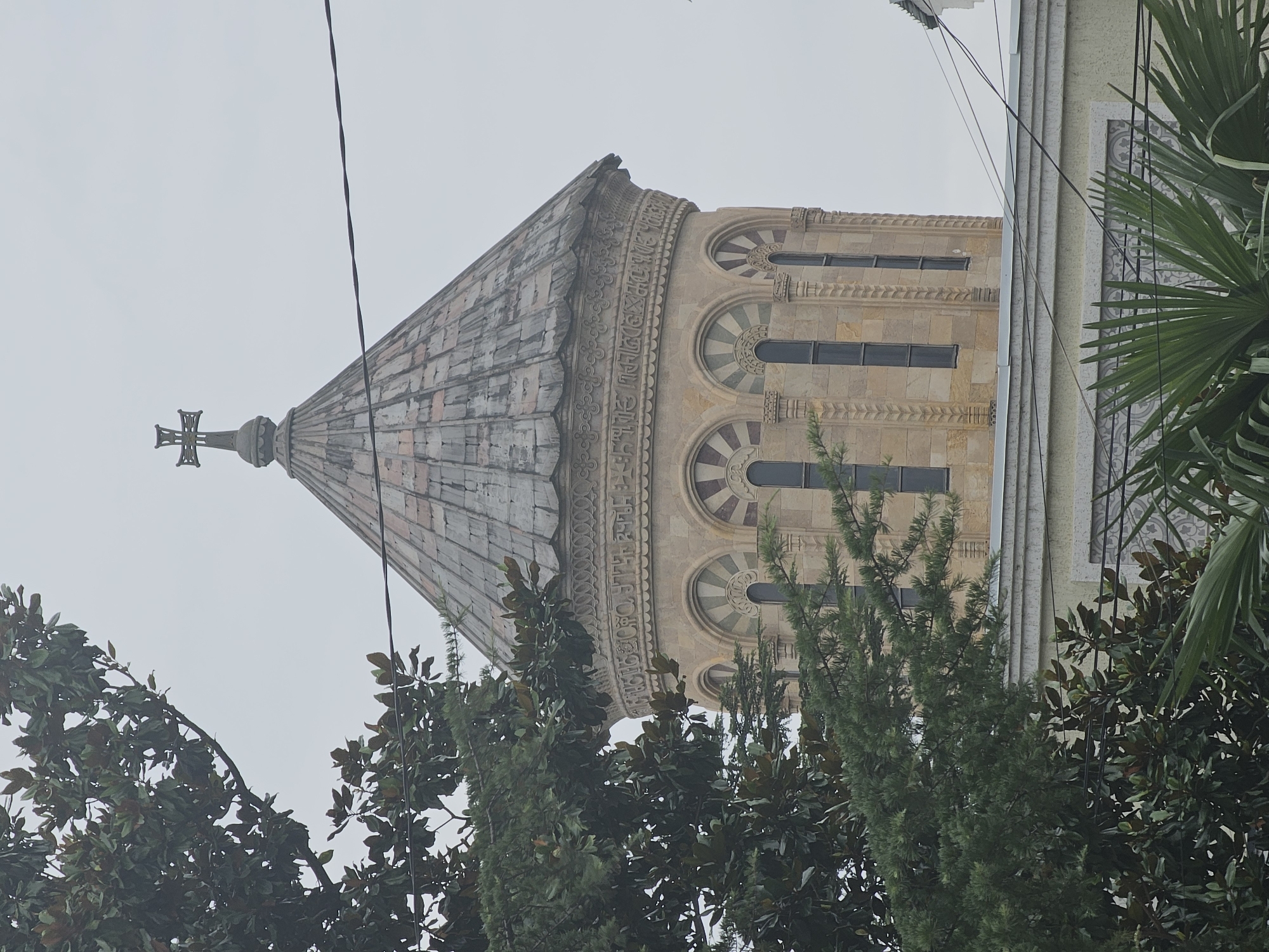 The image shows the upper portion of a domed church or cathedral. The dome is covered in grey tiles and topped with a cross. The cylindrical base of the dome is constructed of light brown stone and features a decorative band of inscribed text and a row of arched windows. Trees with dark green leaves partially obscure the structure. The sky is overcast and light grey. Power lines are visible in the foreground, running across the sky. The building appears to be located in Samtredia, Georgia.