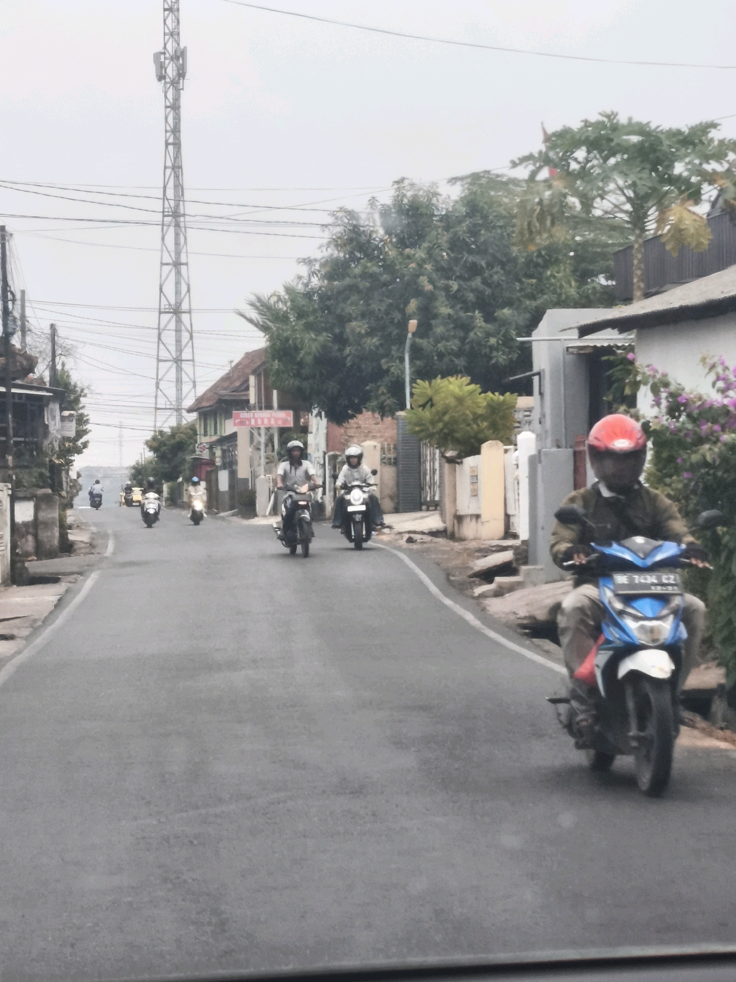 The image depicts a street scene in Bandar Lampung, Indonesia, on an overcast day. Several individuals are riding motorcycles along a paved road that curves to the left. The architecture visible suggests a residential or mixed-use area with buildings of varying styles and sizes lining the street. A prominent cell tower stands tall in the background, indicating a developed urban environment. The road is bordered by sidewalks and some greenery, including trees and bushes. The overall atmosphere is calm, with no apparent urgency or activity beyond the routine movement of traffic.