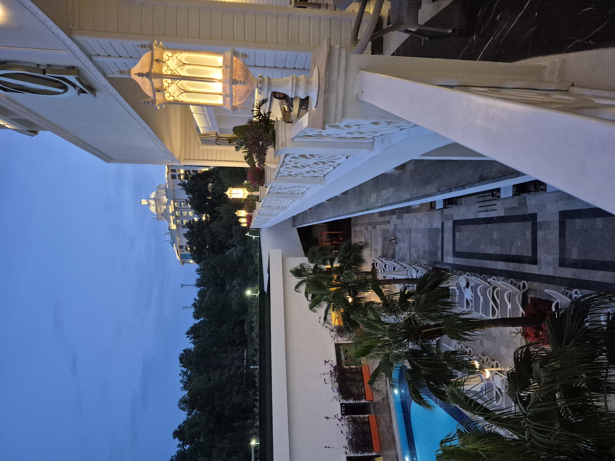 The image depicts an elevated view of a resort or hotel setting in Alanya, Turkey, at dusk. The sky is a gradient of blue, transitioning from light near the horizon to darker shades above. In the distance, a large building, possibly another hotel, is visible with its lights illuminated. A line of trees separates the building from the resort.
The foreground features a balcony with ornate white railings and architectural details. A lighted lantern fixture is prominently displayed, casting a warm glow. Below the balcony, a courtyard area is visible, featuring patterned stone flooring and rows of white lounge chairs, suggesting a relaxation area. A swimming pool is partially visible with bright blue water, surrounded by palm trees. The overall setting indicates a well-maintained and aesthetically pleasing resort environment.