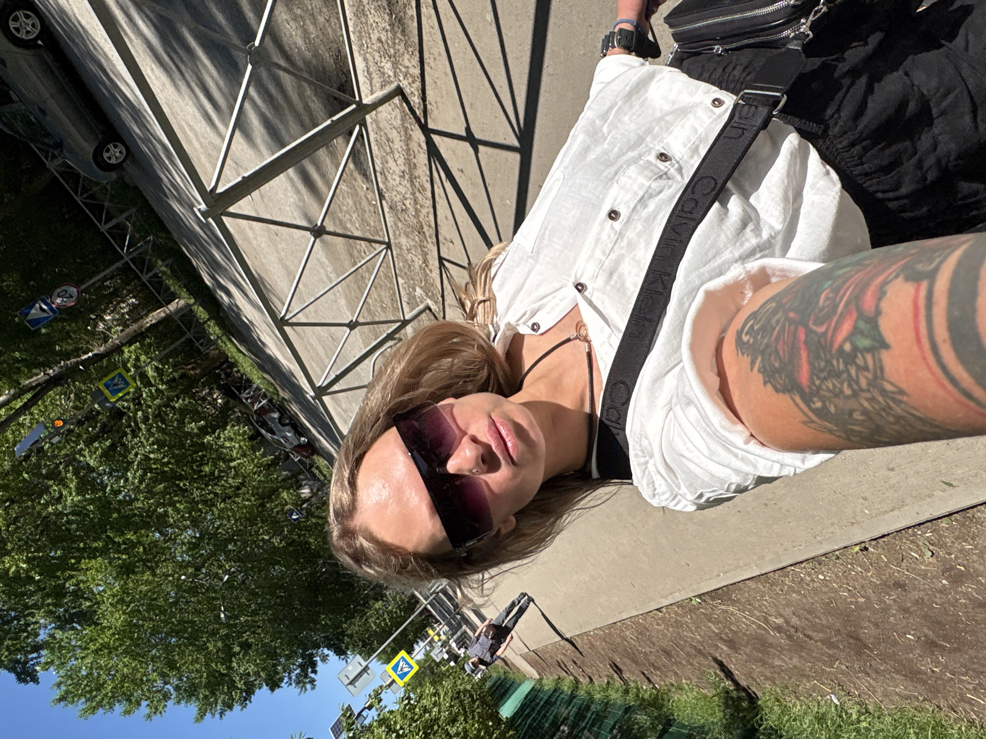 A blonde woman with sunglasses and tattoos on her right arm lies on a sidewalk next to a metal fence. She is wearing a white shirt and a Calvin Klein-branded shoulder strap. The background includes trees, street signs, a car, and a person walking. The scene appears to be in Perm, Russia, based on the street signs.