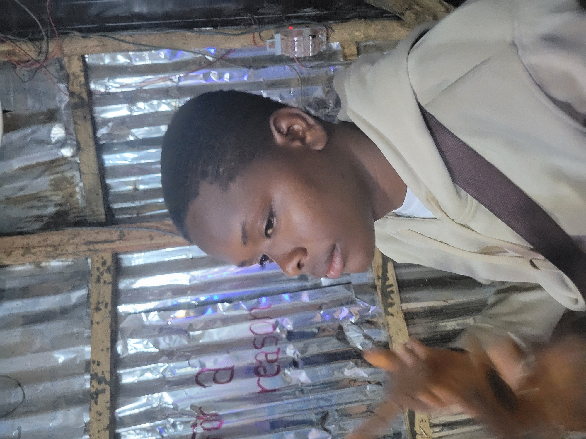 The image shows a young Black person, appearing to be of adolescent or teenage age, in a profile view facing right. They have short, dark hair and are wearing a light-colored hoodie with a darker strap visible across the chest. Their left hand is raised, with fingers extended, partially blurred due to motion. In the background, there is a corrugated metal wall with wooden beams and some electrical wiring. There are also some handwritten words in pink on the metal wall, appearing to read "for a reason." The overall environment suggests an outdoor or semi-enclosed space, possibly in an urban or informal setting. There are no specific landmarks or contextual clues within the image that would allow for definitive identification of the location as Durumi, Nigeria.