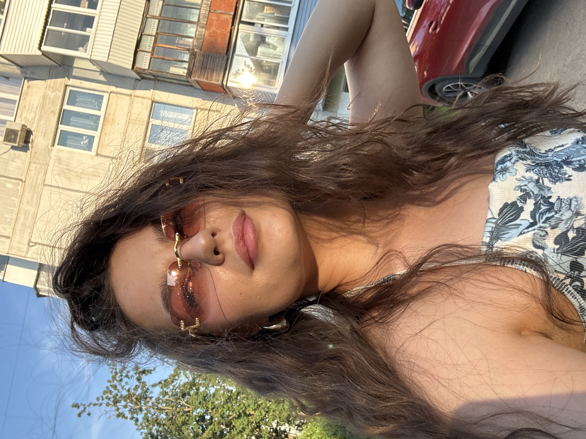A young woman with long, dark, wavy hair is shown from the chest up, taking a selfie outdoors. She is wearing stylish pink-tinted sunglasses with gold accents and a patterned top. Her right arm is raised and behind her head, with her left hand touching her hair. She is looking towards the camera with a neutral expression. The background consists of a multi-story apartment building with balconies and windows, a green tree on the left, and a red car parked on the right. The lighting suggests it is late afternoon or early evening, with the sun casting a warm glow on the woman's face and shoulders. The environment appears to be an urban residential area in Chaikovsky, Russia.