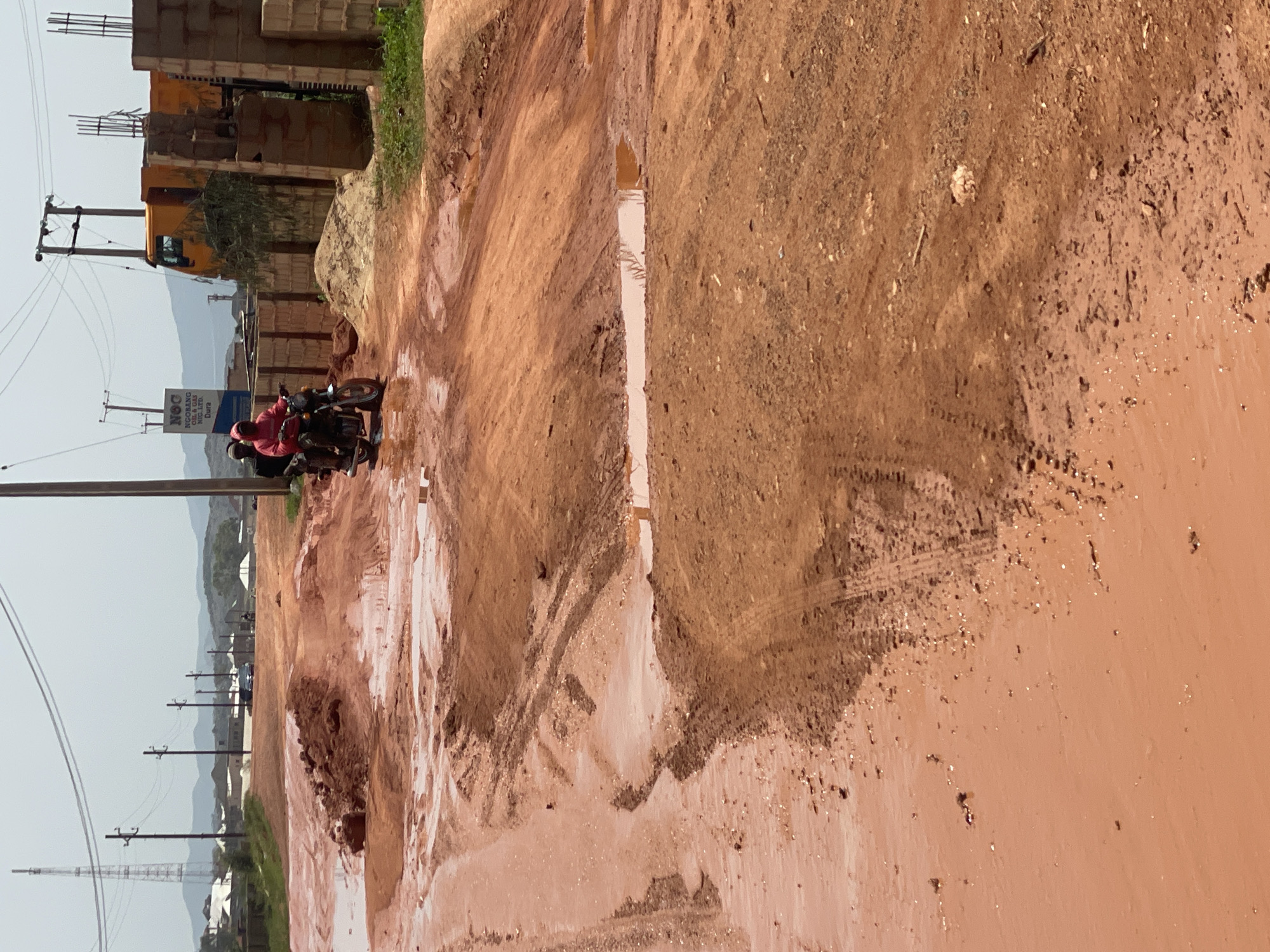 The image depicts a red dirt road in the city of Jos, Nigeria, characterized by significant mud and puddles. Two individuals are riding a motorcycle, with the driver wearing a red hooded sweatshirt and the passenger in black. To the right, construction is evident, with brick walls and rebar visible, along with a yellow dump truck. In the background, distant hills and buildings suggest a semi-urban or developing area. The overall scene captures the challenging road conditions and ongoing construction in Jos.
