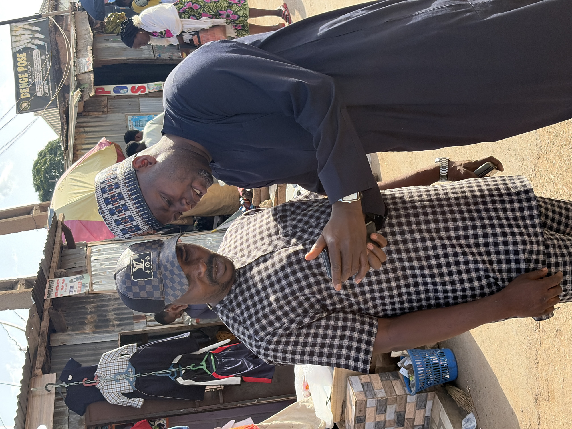 Two men are interacting in a public market area, likely in Gura Top, Nigeria. One man, wearing a blue gown and a patterned cap, leans down towards the other man, who is seated. The seated man wears a checkered shirt, a "LV" monogrammed baseball cap, and holds a cell phone in his hands. They appear to be in conversation, potentially showing affection, as the standing man has his hand on the seated man's back. The background includes market stalls made of corrugated metal, with clothing for sale hanging on a rack. There is a sign for "DENGE POSE NAILS SALOON." A blue plastic basket sits on the ground next to a tiled structure. Other people are visible in the background, suggesting a busy market scene.
