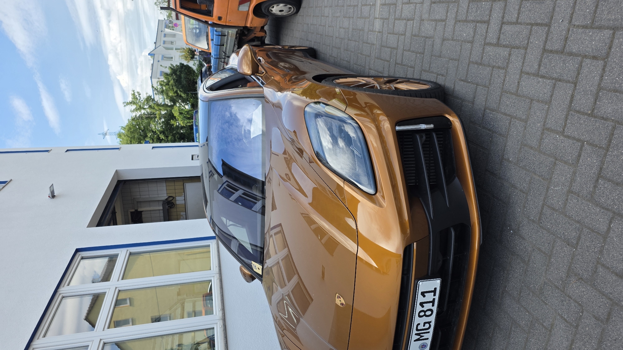 A close-up, low-angle shot captures the front and side of a shiny, copper-colored sports car parked on a cobblestone pavement. The car is the primary subject, with its sleek lines and metallic finish dominating the frame. Reflections of a bright blue sky with white clouds are visible on the car's windshield, and the building to the left. The building is white with several windows and a dark opening. To the far left, a portion of another car, possibly orange, is visible. A license plate on the sports car reads "MG 811". The scene appears to be outdoors during the daytime, under fair weather conditions.