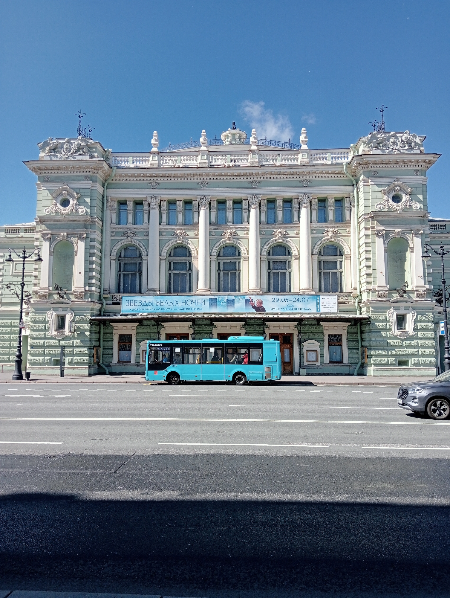 The image captures a bright, sunny day in Saint Petersburg, Russia, with a focus on the Mariinsky Theatre. A light blue public transport bus is prominently displayed on the street in front of the theatre's facade. The theatre itself is an imposing, light green building with ornate architectural details, including columns, arches, and decorative elements. A banner is visible across the theatre's entrance, advertising "ЗВЕЗДЫ БЕЛЫХ НОЧЕЙ" (Stars of the White Nights), with dates 29.05-24.07 and "XXXIV МУЗЫКАЛЬНЫЙ ФЕСТИВАЛЬ" (XXXIV Musical Festival). A dark-colored car is parked on the right side of the street, partially visible. The street is paved and marked with white lines. A lamppost stands on the left side of the image, near the theatre. The sky is a clear, bright blue, indicating fair weather. The scene appears to be a still, everyday moment in the city.