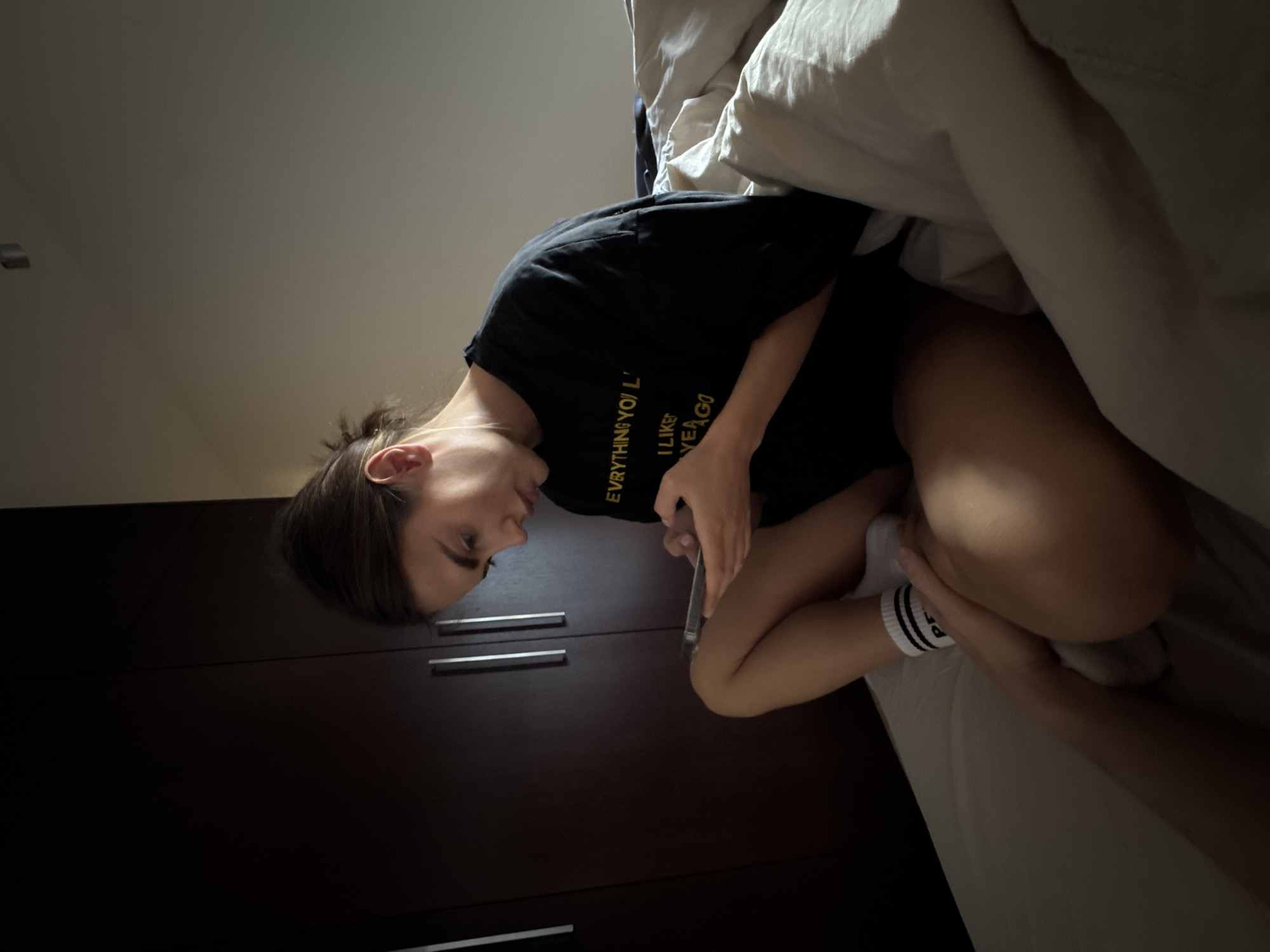 A person with shoulder-length brown hair is sitting on a bed with white linens in an indoor space. The person is wearing a black t-shirt with gold lettering and white socks with black stripes. They are looking down and holding a mobile phone. Behind the person is a dark-colored, possibly wooden, dresser with metal handles. The location is unknown, with no indications of being in Warsaw, Poland.