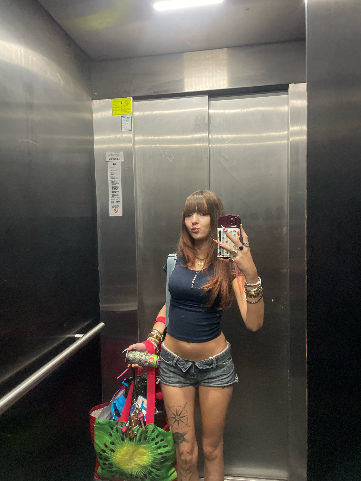 The image depicts a young woman taking a selfie in an elevator. She is wearing a dark blue crop top, denim shorts, and various accessories including bracelets, rings, and a necklace. She is holding a can of aerosol and a tote bag with a kiwi design. The elevator appears to be made of stainless steel, with a black door on the right. There are informational stickers on the elevator doors. The location is identified as an elevator in Raanana, Israel.
