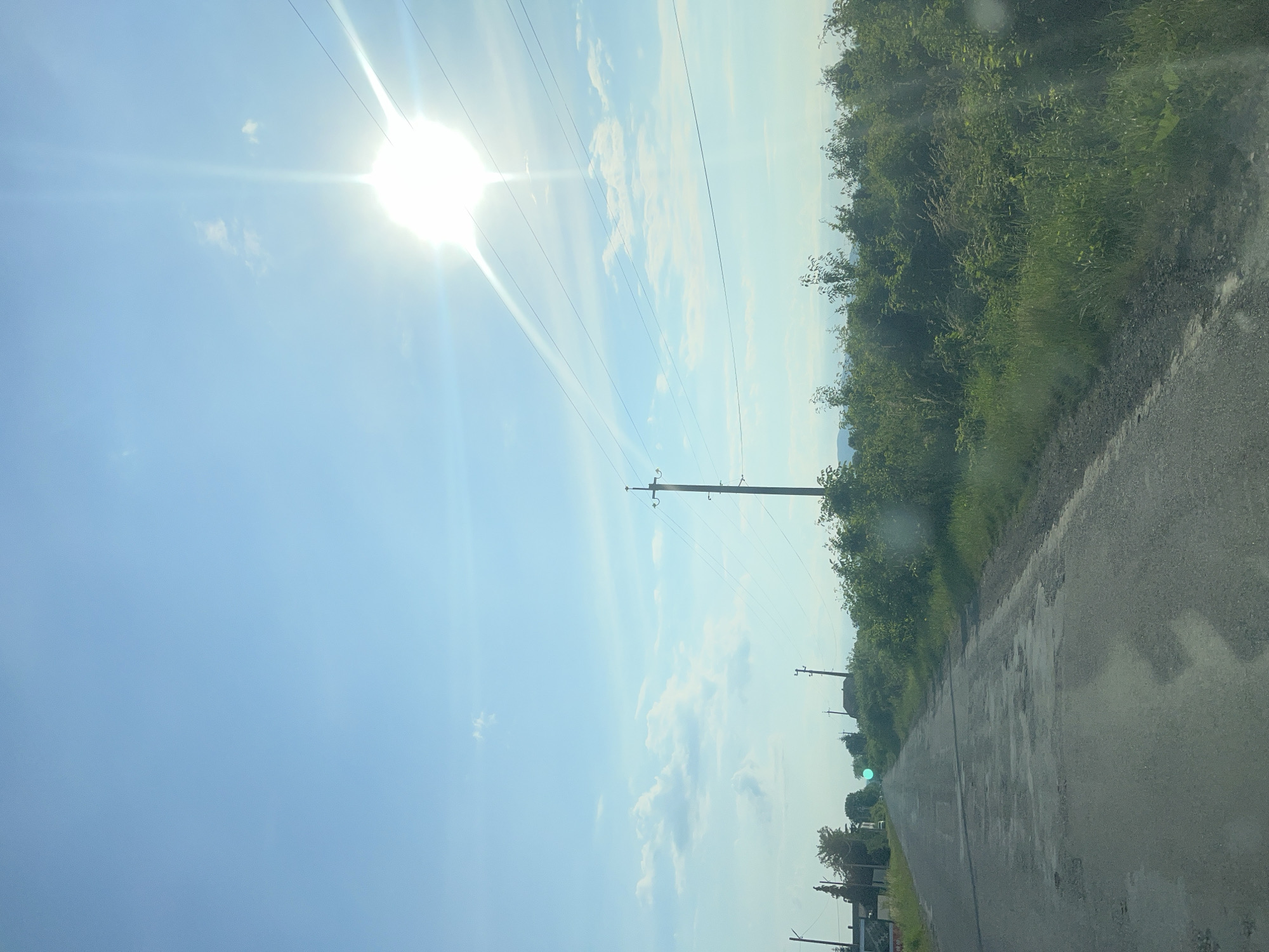 The image shows a rural road in Storozhnytsia, Ukraine, on a sunny day. The road is paved and appears to be in disrepair, with several potholes visible. It is lined on both sides by lush green vegetation, including trees and bushes. Power lines and utility poles run along the right side of the road, stretching into the distance. The sky is a clear blue with a few scattered white clouds. The sun is bright and high in the sky, casting a strong glare and lens flare across the upper right portion of the image. The overall scene depicts a typical rural landscape in Ukraine during daylight hours.