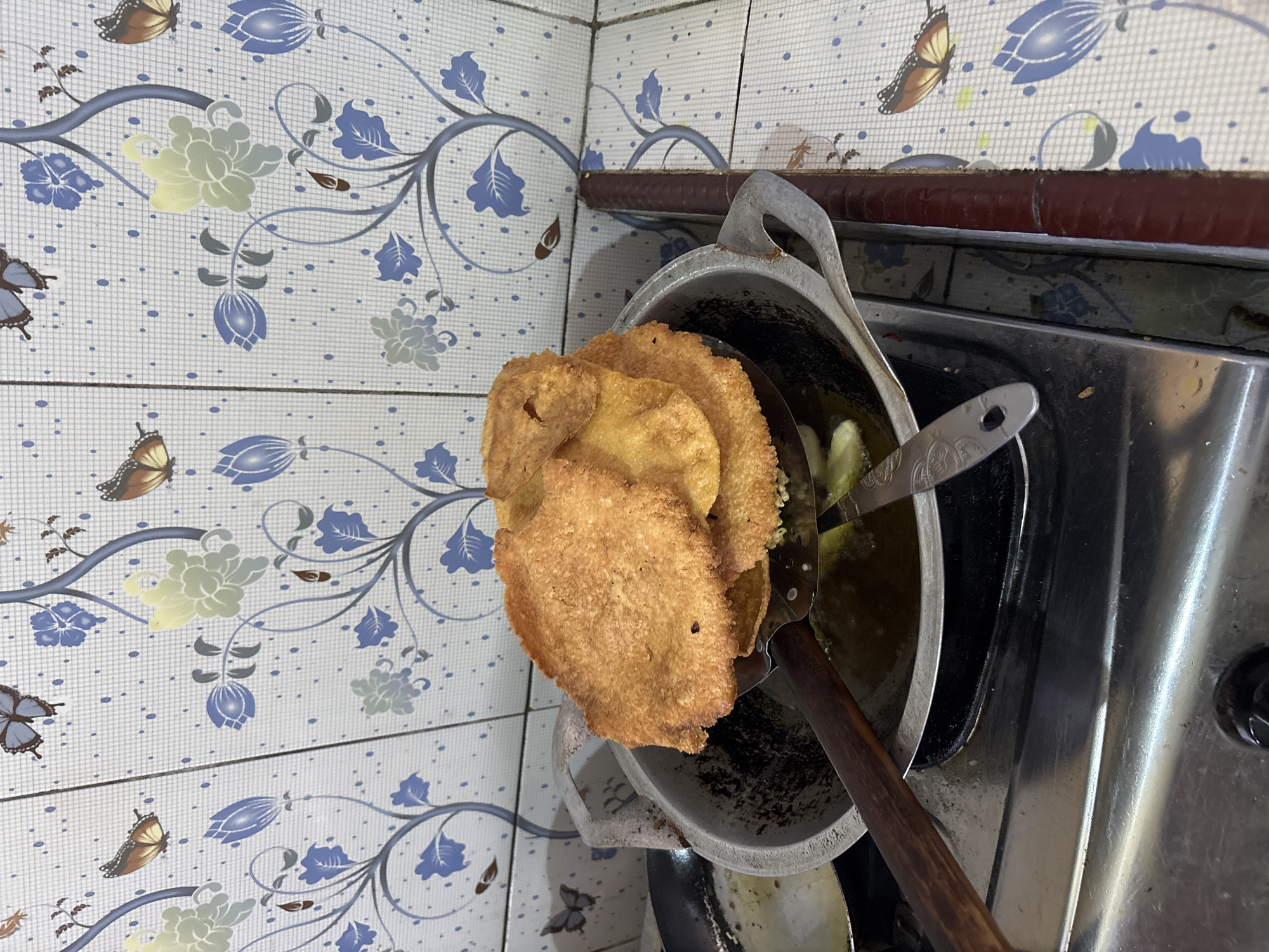 The image depicts a close-up, overhead view of a cooking scene in Tirtomoyo, Indonesia. The focus is on a metal frying pan containing oil, where several golden-brown, crispy fried snacks are being lifted out with a slotted metal spatula that has a wooden handle. The snacks appear flat and irregularly shaped. Behind the pan, a tiled backsplash with a blue and white floral and butterfly pattern is visible. The cooking is taking place on a stainless steel stovetop. The overall impression is of a home kitchen environment where food is being prepared.