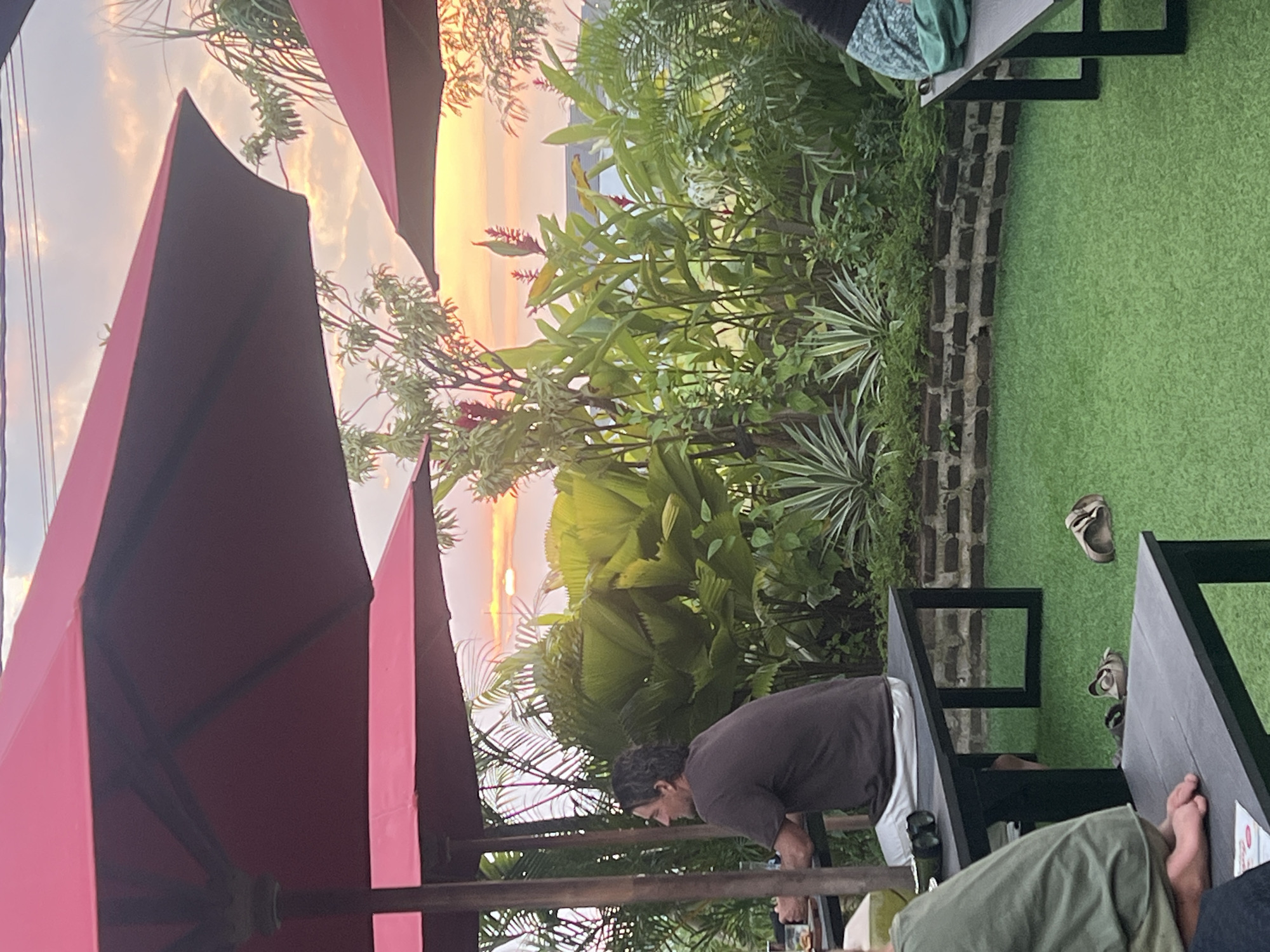 The image depicts an outdoor seating area, possibly a restaurant or cafe, with lush vegetation and a sunset visible in the background. Three large red umbrellas provide shade over black picnic-style tables and benches. On one bench, a man in a dark t-shirt and white pants sits facing left, appearing to be in conversation or observing something outside the frame. Two pairs of sandals are casually placed on the artificial green turf flooring. The setting suggests a relaxed, tropical ambiance, characteristic of locations in or near cities like Kediri, Indonesia, known for their warm climate and vibrant plant life. The lighting indicates it is late afternoon or early evening, with the sun casting a warm glow.
