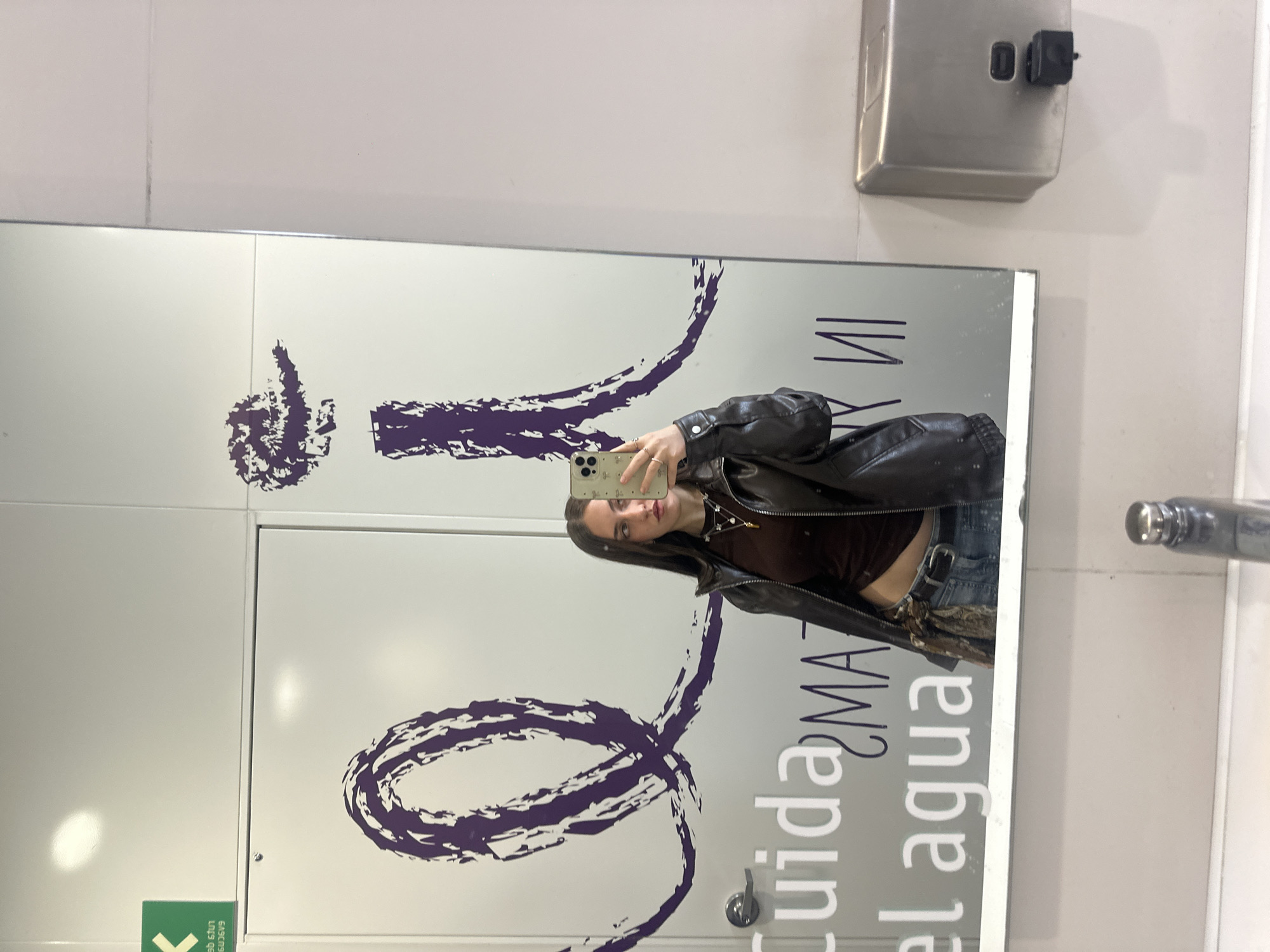 A young woman is captured in a mirror selfie, her reflection appearing in a vertical orientation as if lying down, though she is likely standing. She holds a light-colored smartphone with multiple cameras up to take the photo, partially obscuring her face, but her eyes are visible as she looks towards the screen. She has long, dark hair and a neutral expression.
She is wearing a dark brown, fitted top, a black faux leather jacket, and blue jeans, the waistband of which is visible. Her left hand, adorned with several rings, supports the phone. A delicate necklace with small charms is visible around her neck.
The setting appears to be an indoor space, likely a public restroom or changing area, characterized by light-colored, possibly tiled walls. A stainless steel wall-mounted soap dispenser with a black nozzle is visible above the mirror to the right. Part of a silver sink faucet can be seen on the far right.
The large mirror features prominent purple, abstract, brushstroke-like designs or decals. Reflected on the mirror's surface, in mirrored and upside-down text, are the Spanish words "Cuida el agua," meaning "Conserve water" or "Take care of the water," suggesting a public facility in a Spanish-speaking country. A green sign with indecipherable white text, also reflected and upside down, is partially visible in the lower left corner of the mirror. The overall lighting is bright and artificial.