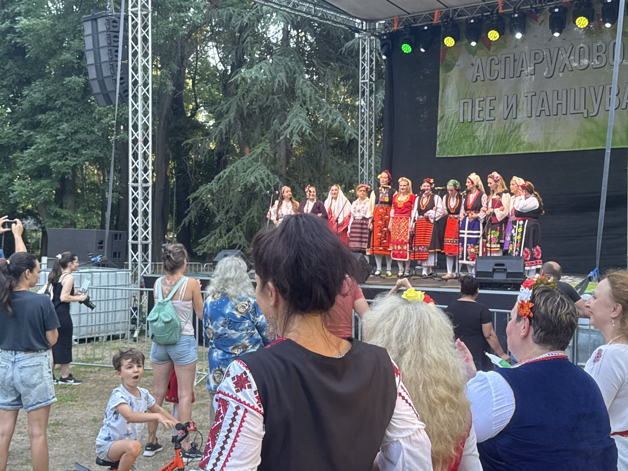 This image captures an outdoor performance on a stage, likely a cultural event, in Varna, Bulgaria. A group of individuals, primarily women dressed in traditional folk costumes, are gathered on the stage. Their attire is colorful and features intricate embroidery and headwear adorned with flowers. They appear to be singing or performing, with some holding microphones.
In the foreground and surrounding area, a crowd of spectators is visible, observing the performance. Among them are men, women, and a young boy on a bicycle, suggesting a family-friendly event. Some audience members are also wearing elements of traditional clothing, such as embroidered vests and floral headpieces, indicating a shared cultural celebration.
The setting is outdoors, with trees providing a backdrop to the stage and surrounding area. The lighting suggests it is late afternoon or early evening, with stage lights illuminating the performers. The overall mood seems festive and community-oriented.
Behind the performers, a large banner is displayed with text. The visible text appears to be in Cyrillic script and reads "АСПАРУХОВО ПЕЕ И ТАНЦУВА," which translates to "Asparuhovo Sings and Dances." This further confirms the nature of the event as a local cultural festival. Other elements in the scene include stage equipment such as speakers and scaffolding, as well as portable barriers separating the audience from the performance area.