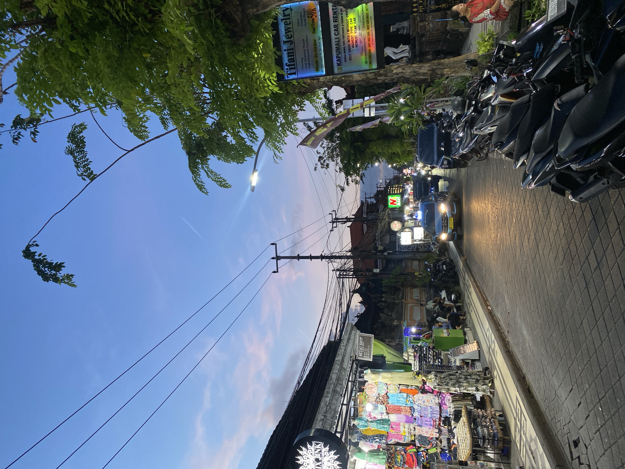 This image captures a bustling street scene in Kuta, Indonesia, during dusk or early evening. The sky, occupying the upper-left portion, displays a gradient of blue fading into soft pinks and purples, indicating the transition from day to night. Lush green tree foliage extends from the left, overhanging the street.
Along the left side of the road, several commercial signs are prominently displayed. A blue sign for "Tifani Jewelry" advertises "Piercing," "Full Diamond Watch," "Full Diamond Necklace," "Silver," "Titanium," and "Gold Plated" services, noting "Made To Order" items "Start from 50k" at "Jln. Raya Legian No. 28A Kuta-Bali." Adjacent to it, a sign for "KAPUMALI CAR RENTAL" lists "SELF DRIVE WITH / WITHOUT DRIVE" options for "HIGHEST TYPE CAR" and "NEWEST YEAR CAR," alongside several contact numbers and social media handles. A bright street lamp illuminates this area.
The paved road runs through the mid-right of the image, where a blue car with headlights on drives towards the viewer, followed by other vehicles in the distance. Along the right side of the road, numerous motorcycles are parked in a neat row. Behind them, market stalls, brightly lit and displaying colorful textiles, clothing, and various goods, line the sidewalk. Signs for "KETUT SHOP," "XNK," and "M MART" are visible above these businesses. Several people are gathered near these stalls, engaged in shopping or conversation. One person in a red top is partially visible in the upper-right corner.
Overhead, a complex network of power lines stretches across the sky, connecting utility poles. The scene portrays a vibrant commercial area coming alive with activity as evening descends.