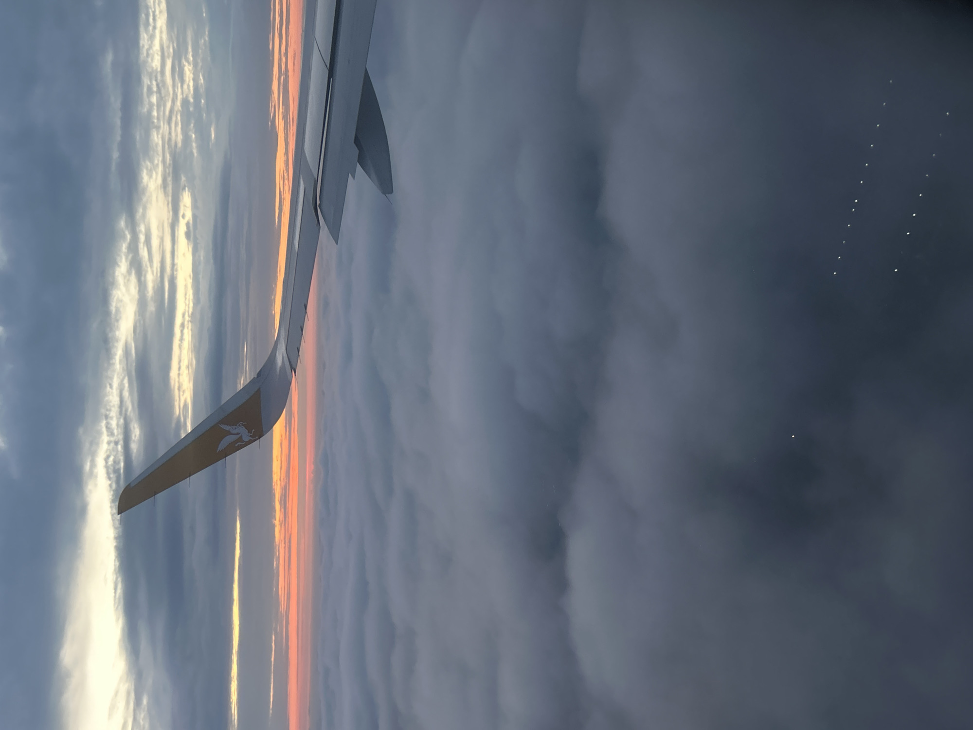 The image shows the wing of an airplane against a backdrop of clouds and a sunset or sunrise. The wing has a logo on it, and there are lights visible on the wing. The scene appears to be taken from inside the airplane, looking out the window. There is no indication of the city of Saint Petersburg, Russia, or any people, subjects, objects, actions, or interactions beyond what is described.