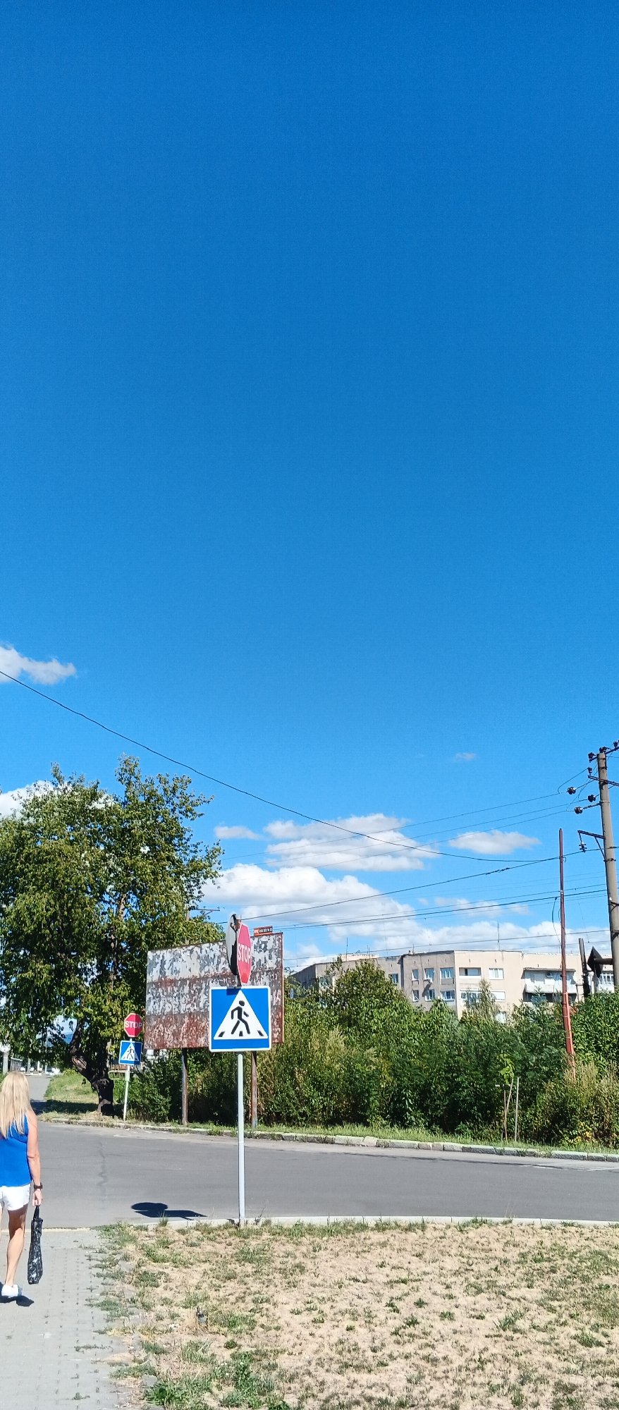 A woman with blonde hair, wearing a blue top and white shorts, walks along a paved sidewalk next to a dry, grassy area. She is carrying a black umbrella. In the background, there is a road with a pedestrian crossing sign and a stop sign on a rusty metal structure. Beyond the road, there are trees, some buildings, and power lines under a clear blue sky with scattered white clouds. The scene appears to be in Mukachevo, Ukraine, based on the context provided.
