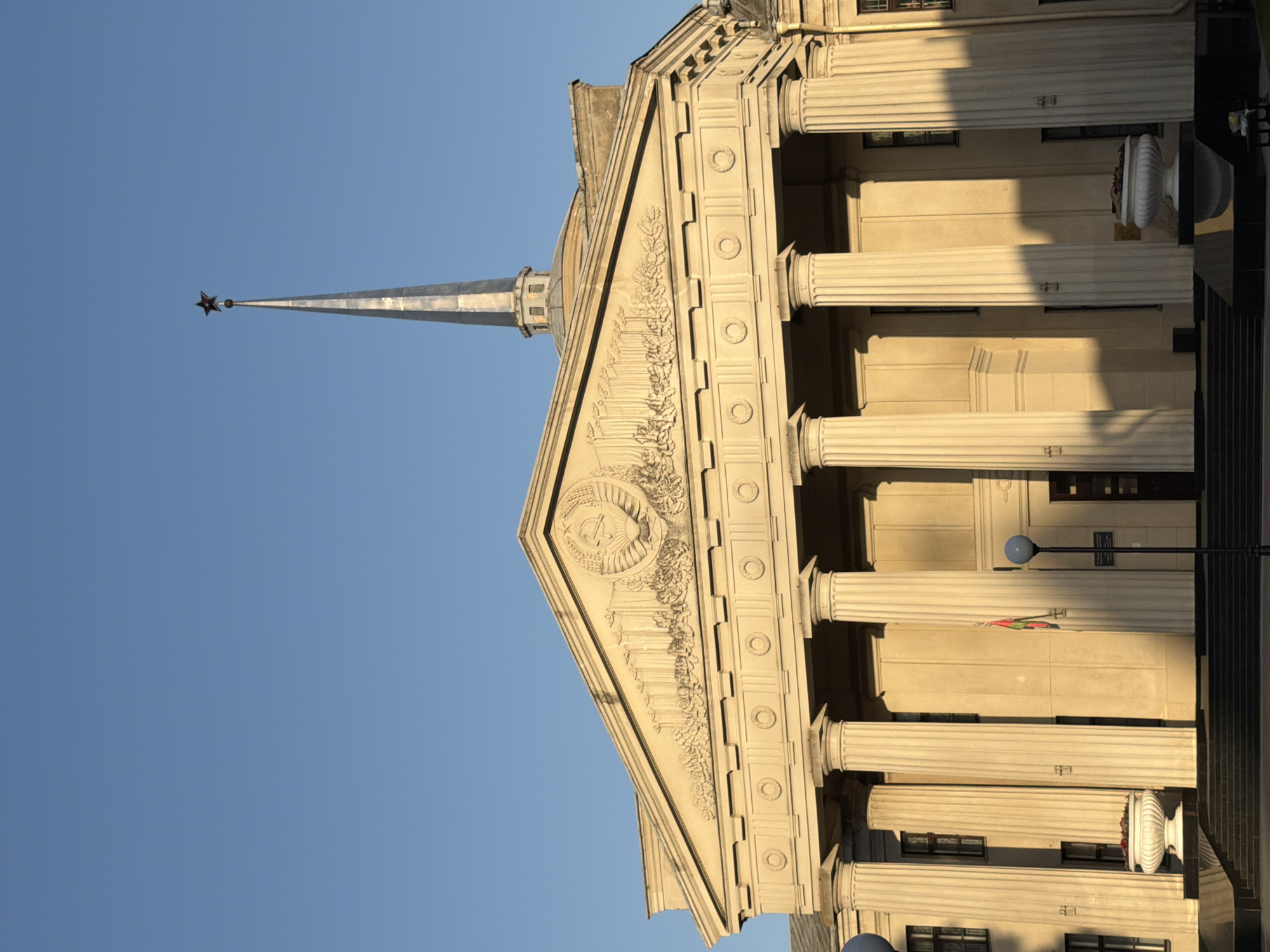 The image shows a neoclassical building with a prominent triangular pediment adorned with a bas-relief emblem of a hammer and sickle, set against a clear blue sky. The building features a colonnade of large, fluted Ionic columns supporting the entablature and pediment. A tall, slender spire topped with a red star rises from the center of the roof structure.
The facade is constructed of light-colored stone, with shadows cast by the late afternoon sun highlighting the architectural details. Below the columns, a dark, tiered staircase leads up to the entrance. Two large, white urns with red flowers are placed on either side of the staircase. A flag, appearing to be the flag of Belarus, is visible hanging from a pole between two of the columns. A street lamp with a spherical light fixture stands to the right of the central section of the building. The overall impression is one of imposing grandeur and governmental significance.