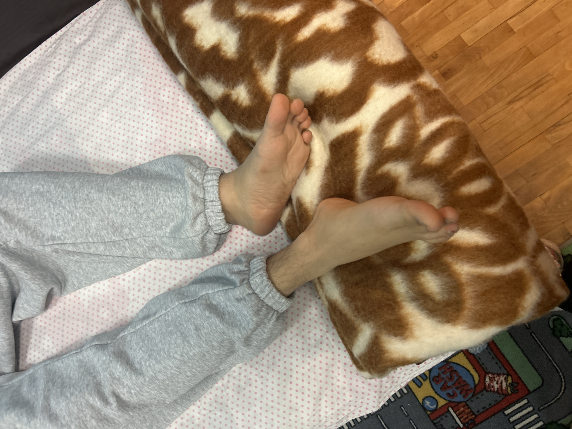 A top-down view shows a person wearing gray sweatpants with bare feet resting on a bed. The bed is covered with a white sheet featuring a pattern of small pink polka dots. Adjacent to the person's feet is a rolled-up blanket with a brown and white animal-like pattern. The setting appears to be indoors. Part of a colorful playmat depicting roads and buildings is visible in the bottom right corner. The location information, Panagyurishte, Bulgaria, cannot be confirmed from the image.