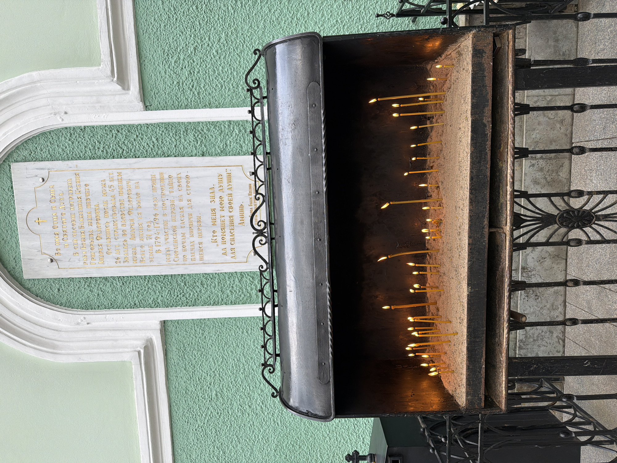 The image shows a candle stand with lit candles outside a green building with a white plaque. The plaque is in Russian and appears to be a memorial inscription. The candle stand is made of dark metal with decorative scrollwork. The candles are yellow and are partially burned. The scene is likely located in Saint Petersburg, Russia, based on the writing on the plaque and the style of architecture.